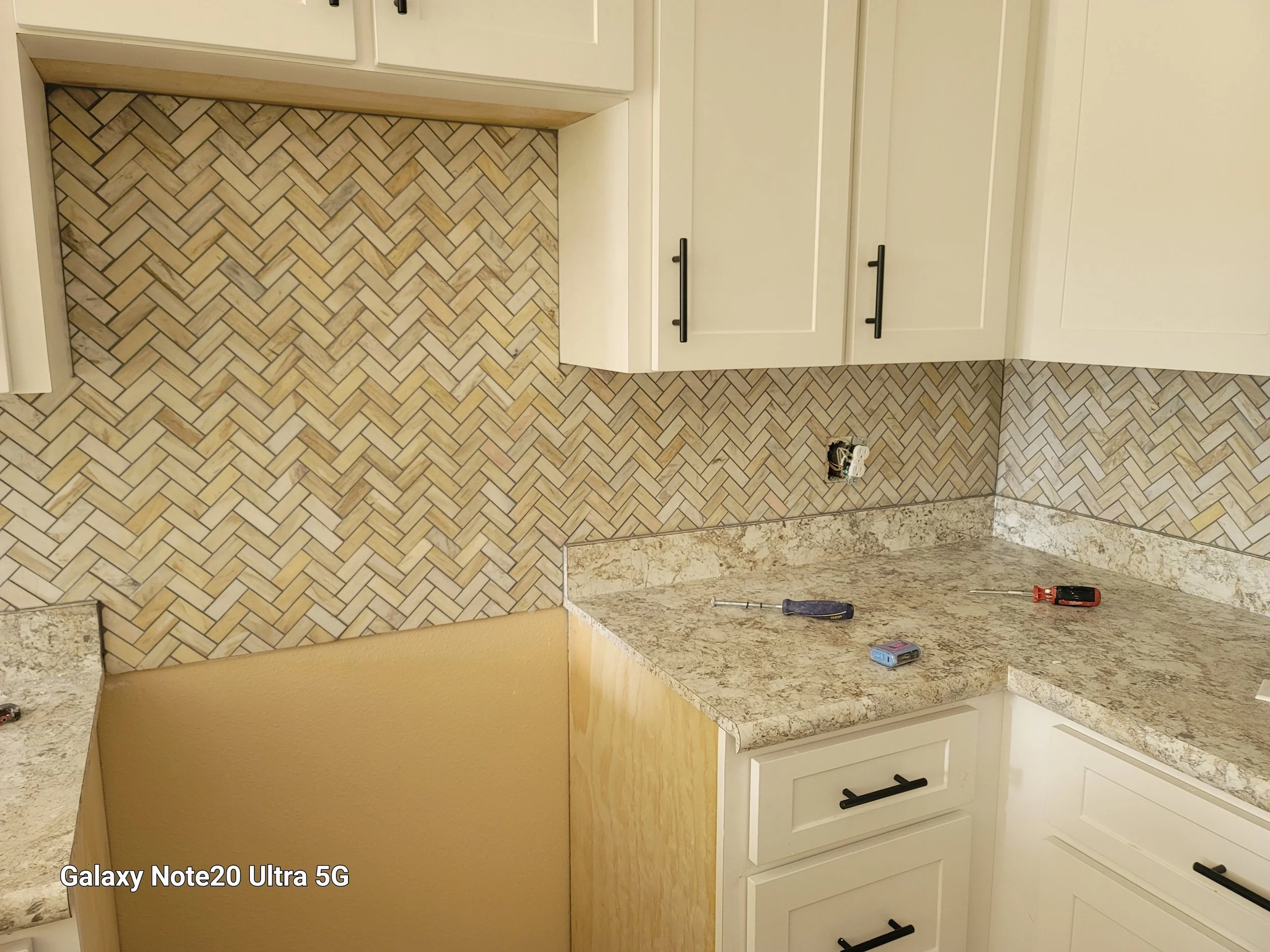 Kitchen with white cabinets, black handles, beige and brown herringbone tile backsplash, granite countertops, and tools including a screwdriver, voltage tester, and power outlet.