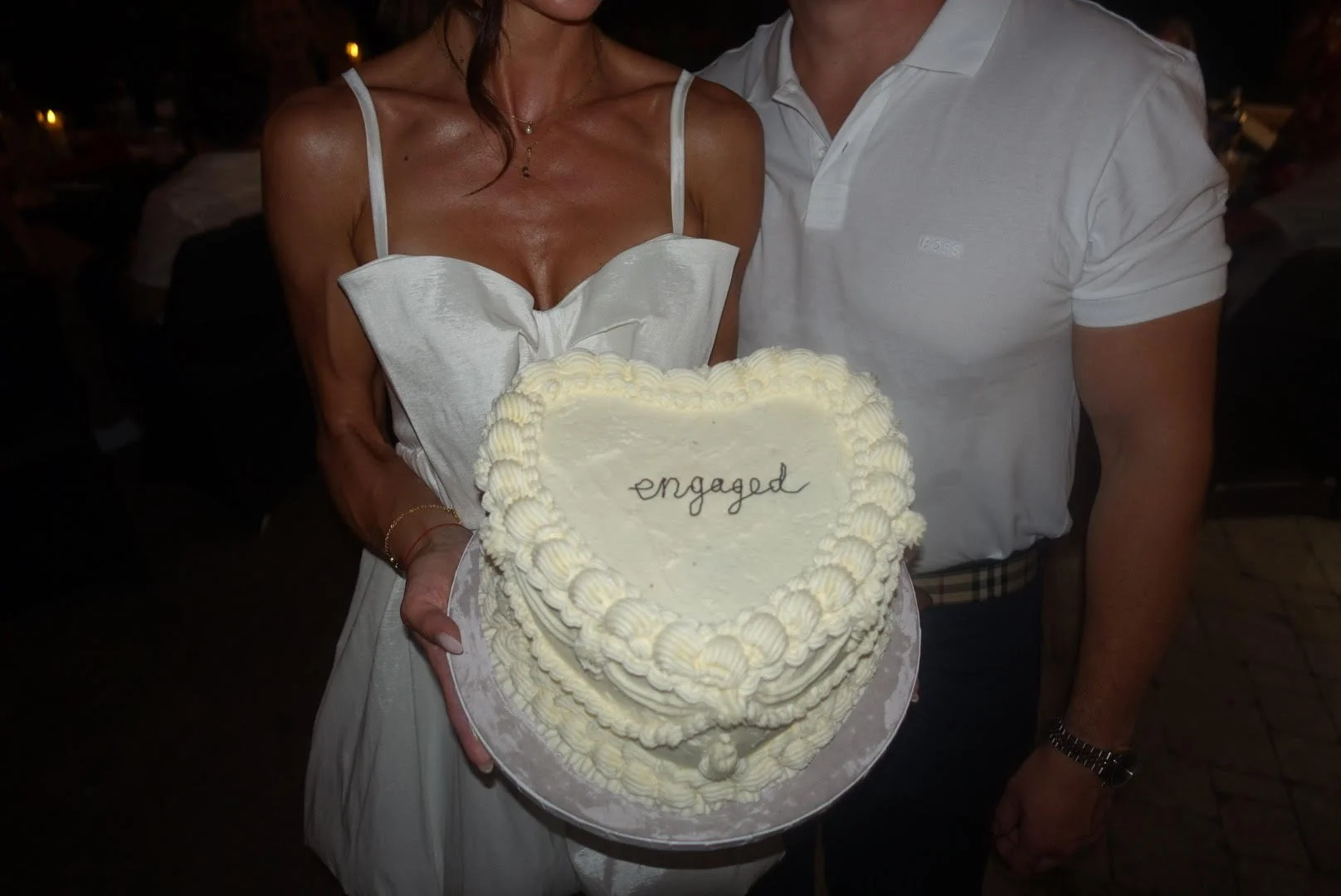 A woman and a man holding a heart-shaped wedding cake with the word 'engaged' written on top.
