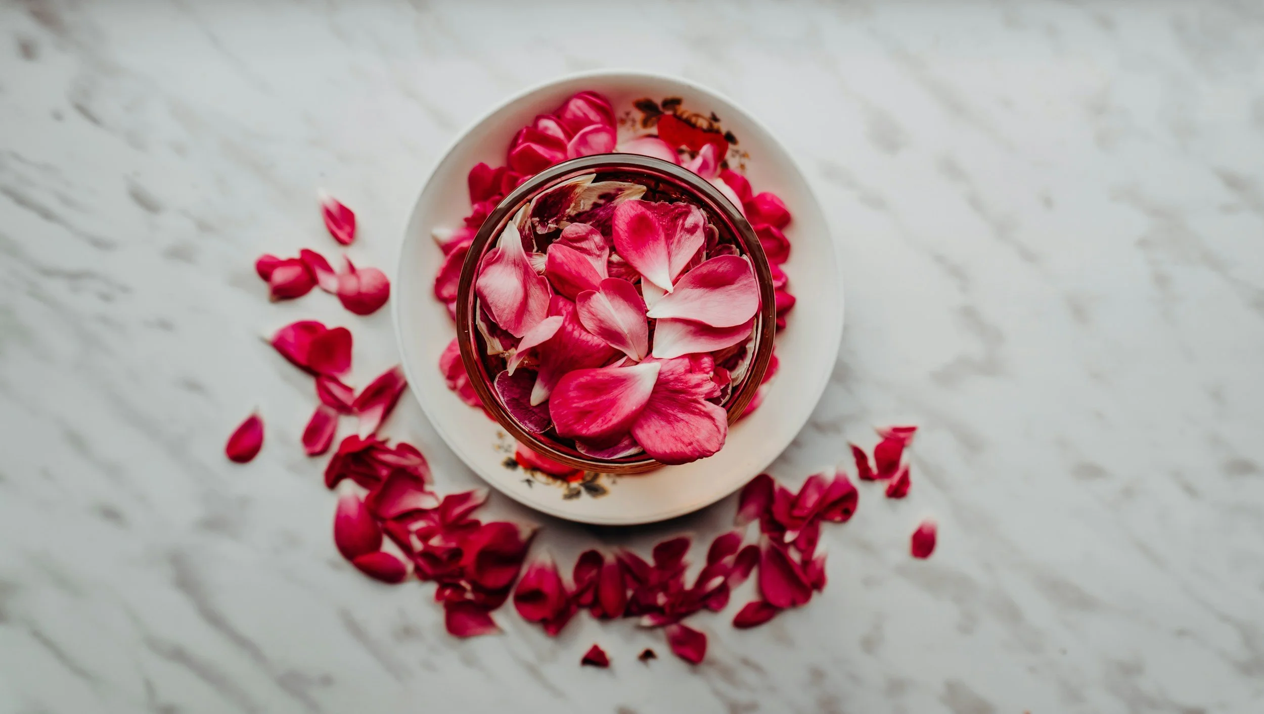 A glass of water filled with rose petals on a white plate, with more scattered petals on a white marble surface.