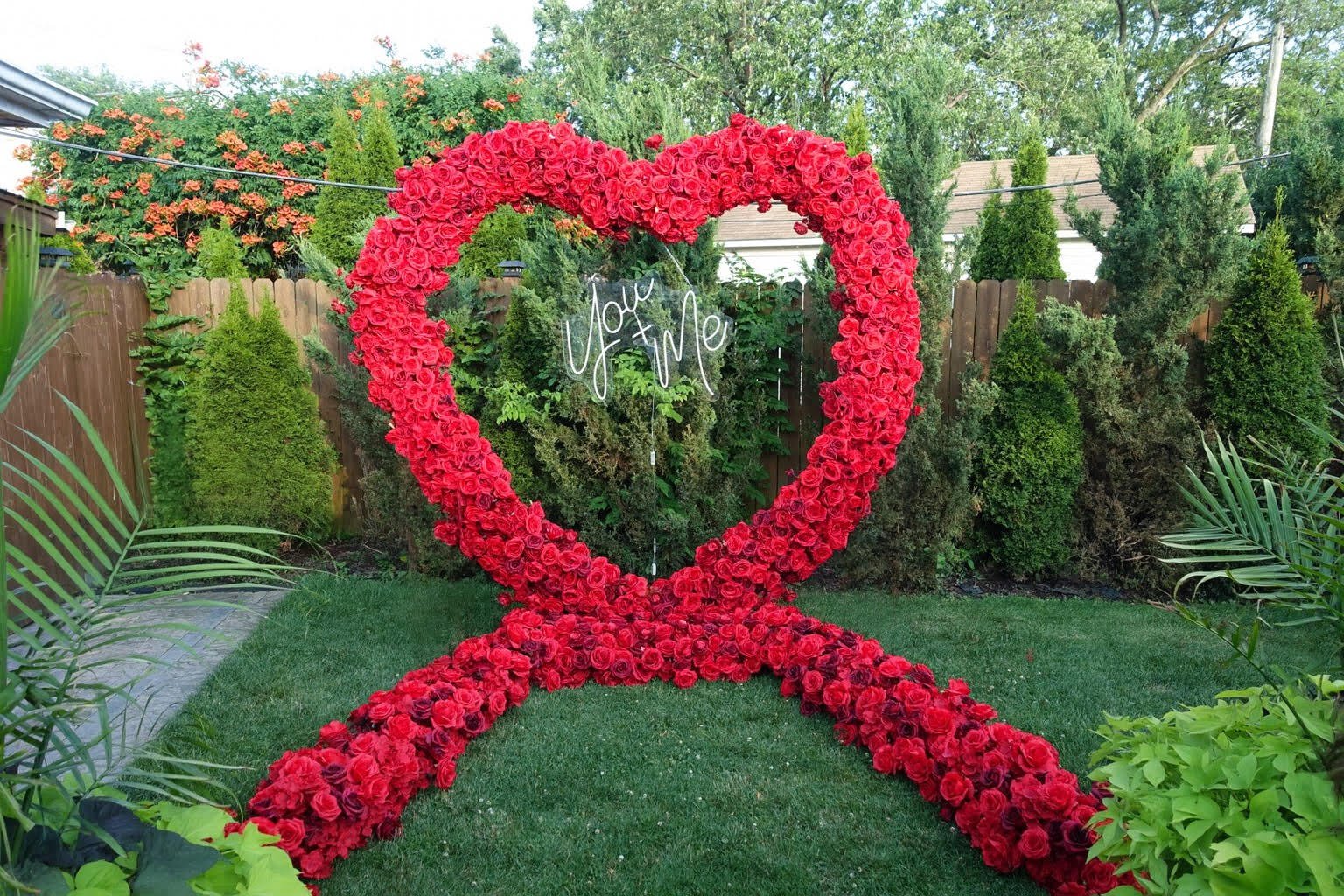 A garden decorated with a large heart-shaped arch made of red roses. In the background, there is a neon sign that reads 'You + Me'. The garden is surrounded by green bushes, trees, and a wooden fence.