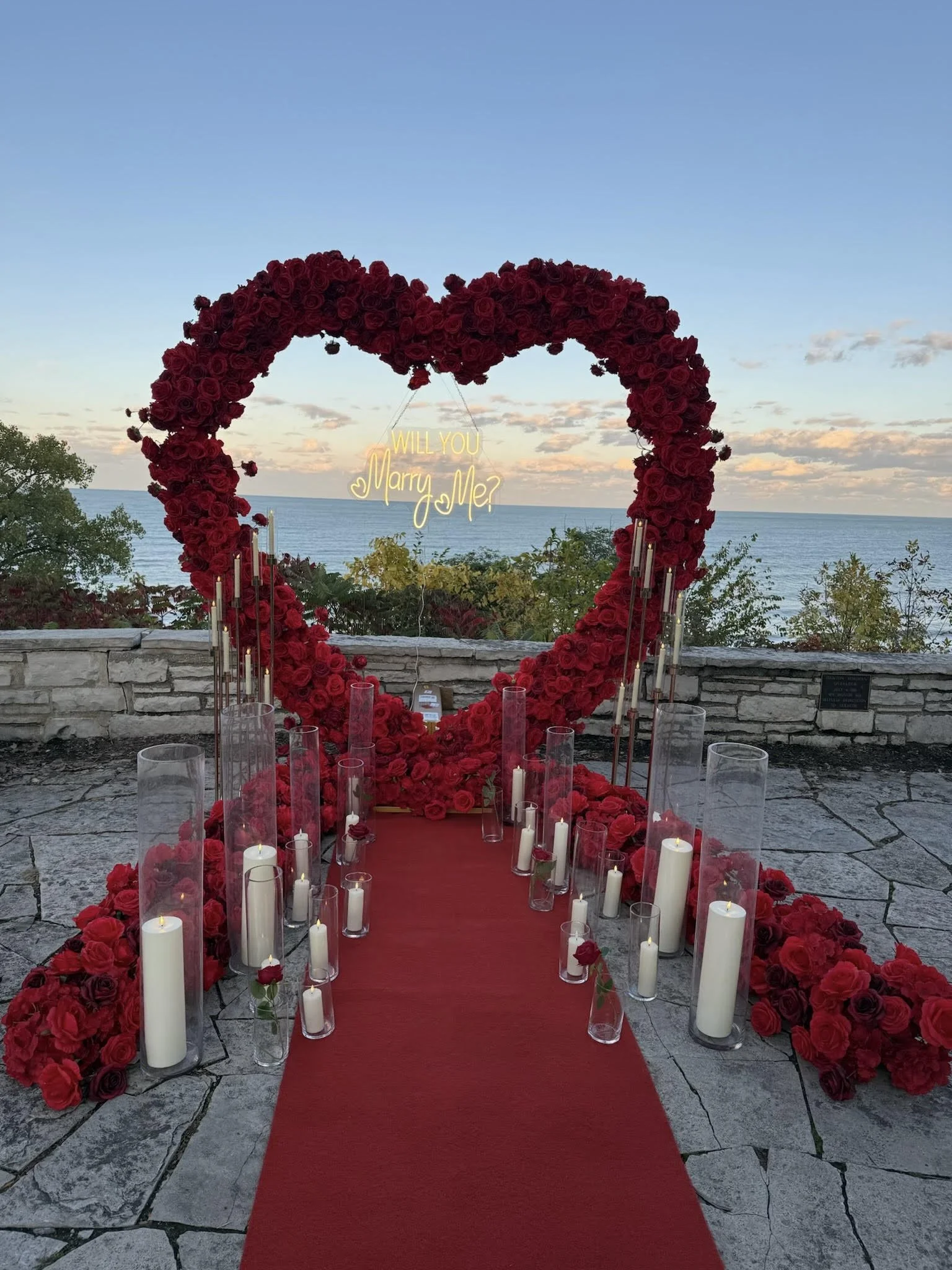 A large heart-shaped floral arch made of red roses with a neon sign inside that reads 'Will you marry me?' overlooking the ocean at sunset. Candles in glass holders line a red carpet leading to the arch.
