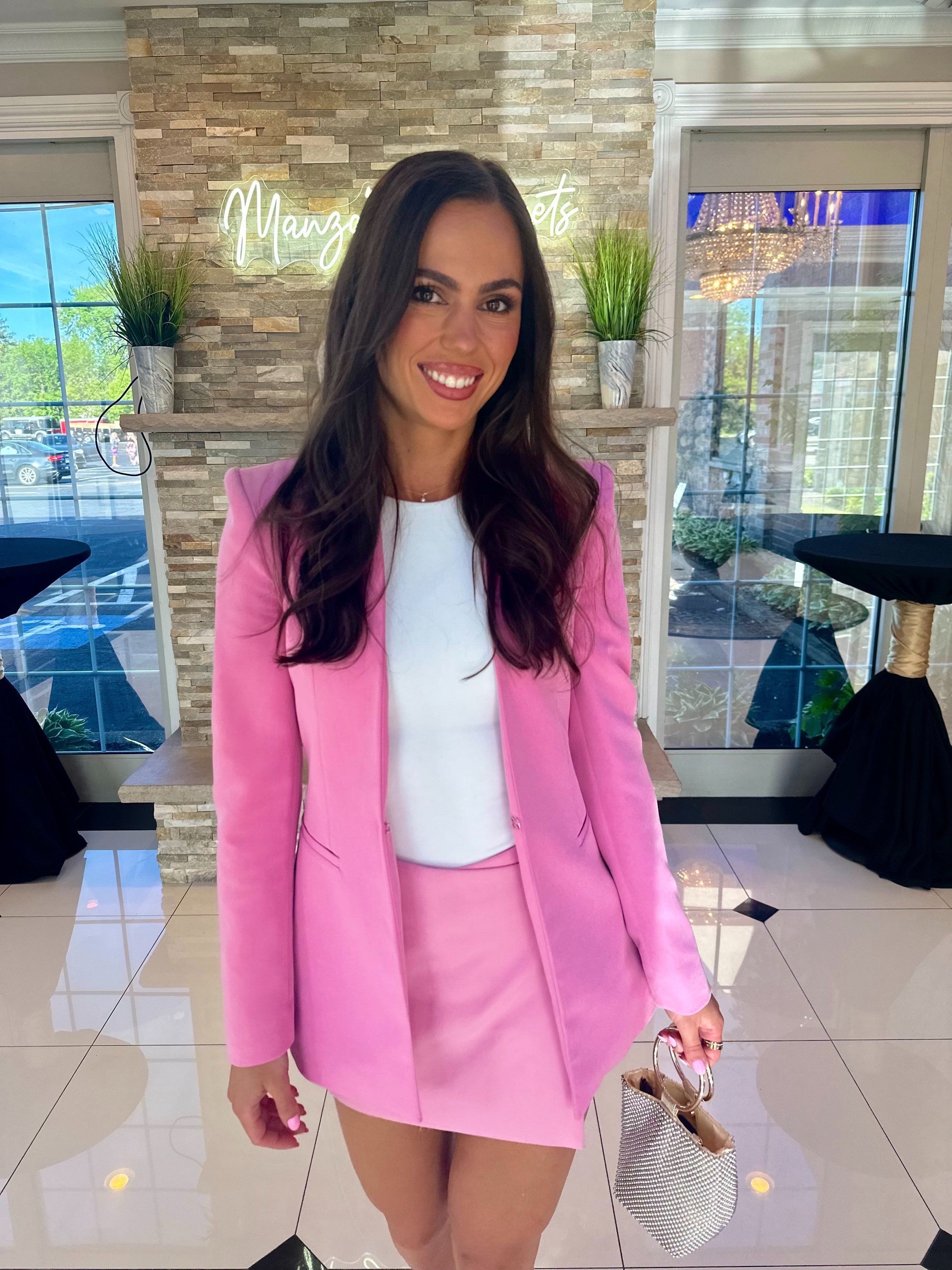 A woman with long dark hair smiling in a pink blazer and skirt, holding a small handbag, standing indoors in front of a stone wall with a sign that reads 'Mango Sets'. The background includes large windows, plants, and furniture.