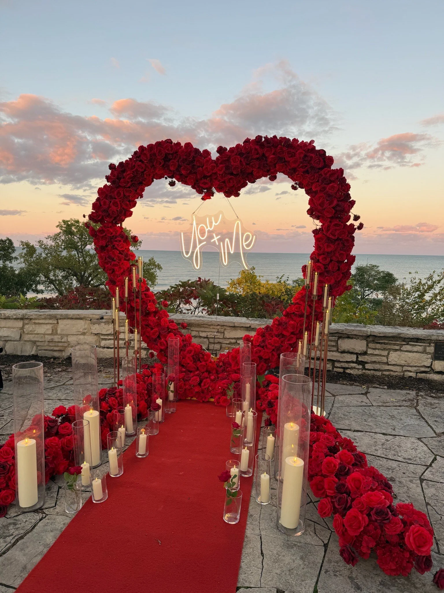 A romantic outdoor wedding aisle with a large heart-shaped floral arch made of red roses, with a neon sign inside reading 'You + Me'. The aisle is lined with candles in glass holders on a red runner, overlooking a sunset view of the ocean and skies with scattered clouds.