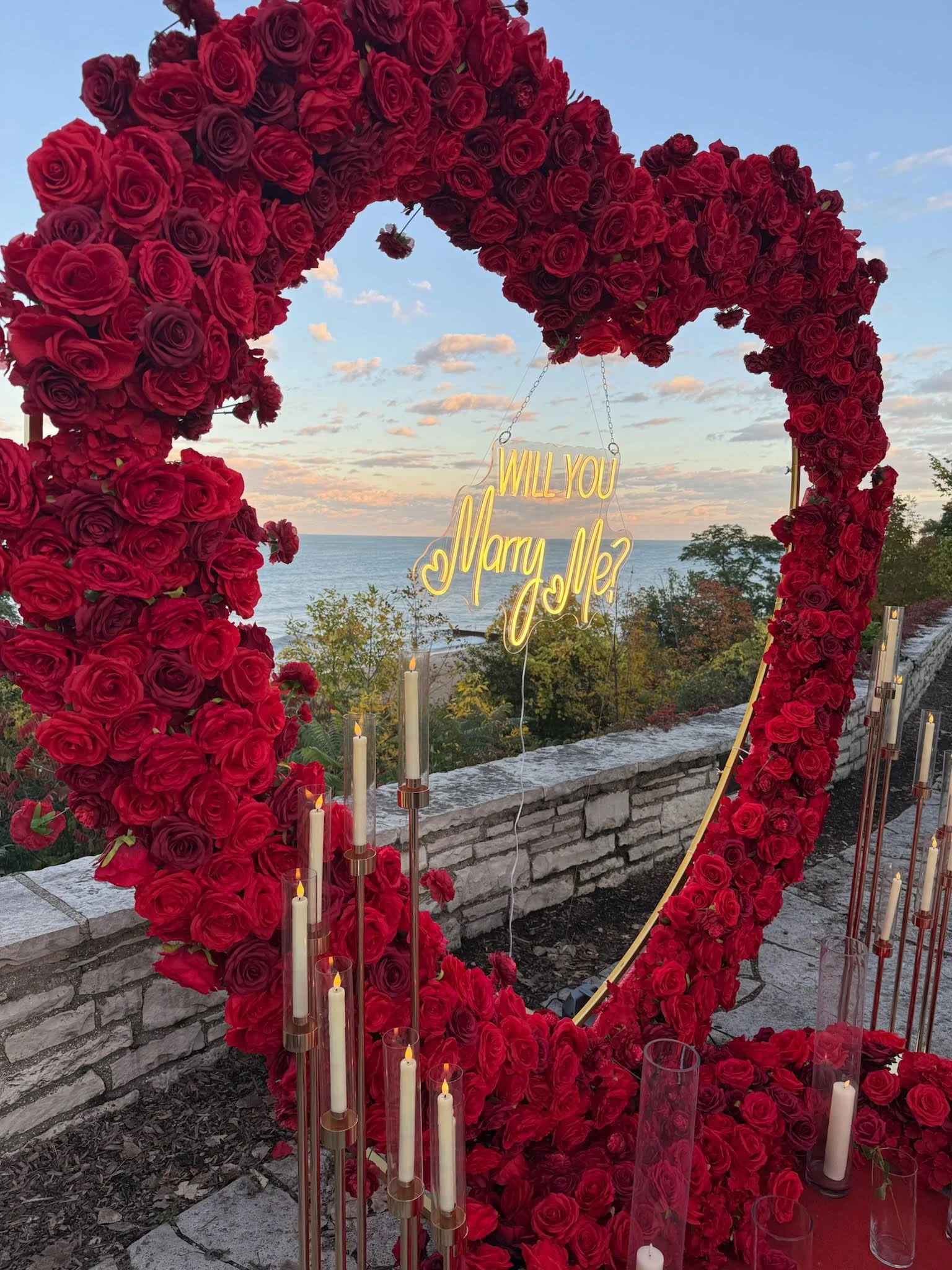 A large heart-shaped arch made of red roses with a neon sign that says 'Will you marry me?' hanging in the center. There are several tall white candles in glass holders at the base of the arch. The background features a scenic view of the ocean and a partly cloudy sky during sunset or sunrise, with trees in the foreground.