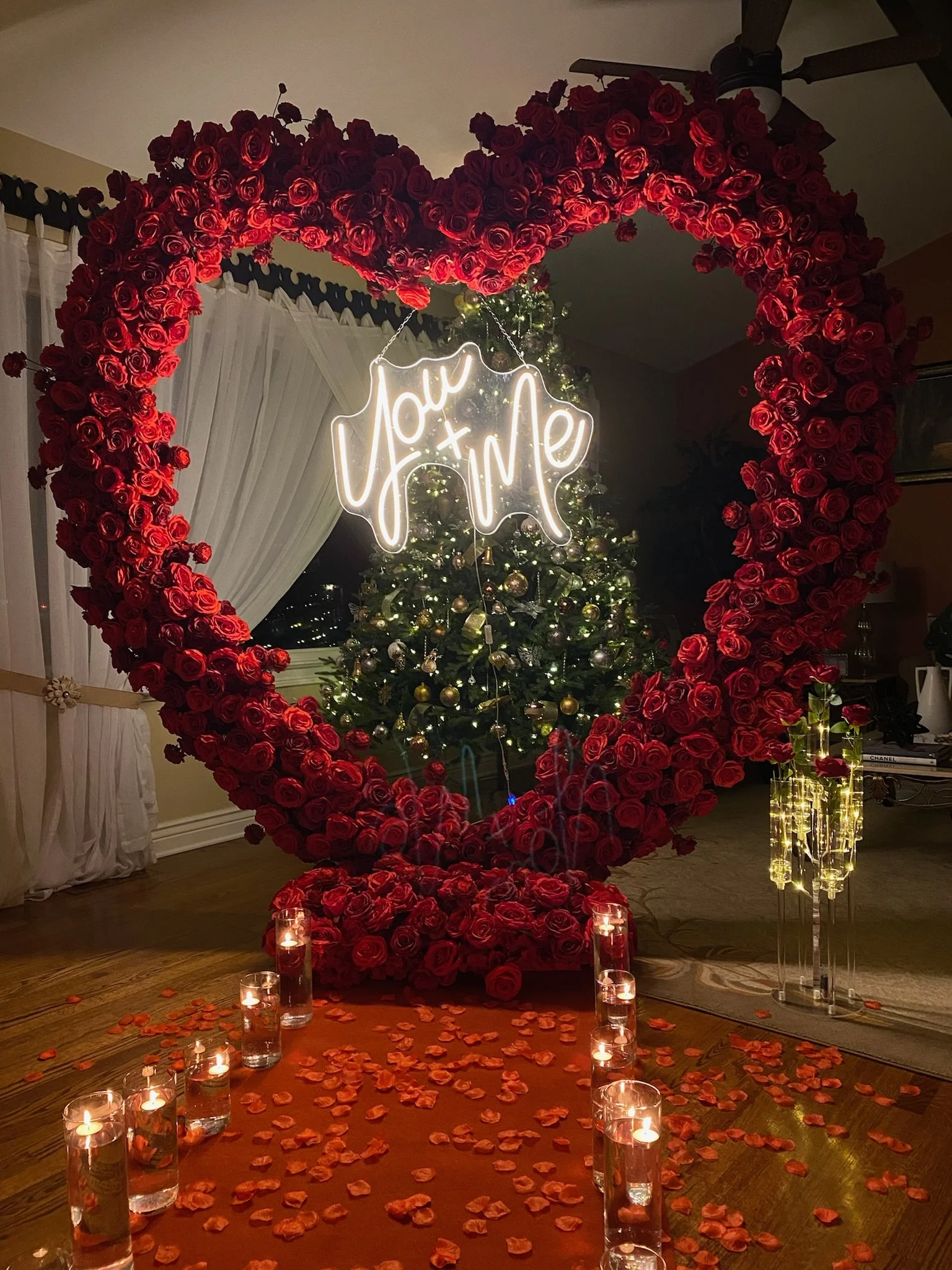 A large heart-shaped frame made of red roses surrounds a glowing sign that says 'You + Me'. In the background, there is a decorated Christmas tree with lights and ornaments, and the scene is set with lit candles and rose petals on the floor, creating a romantic setting.