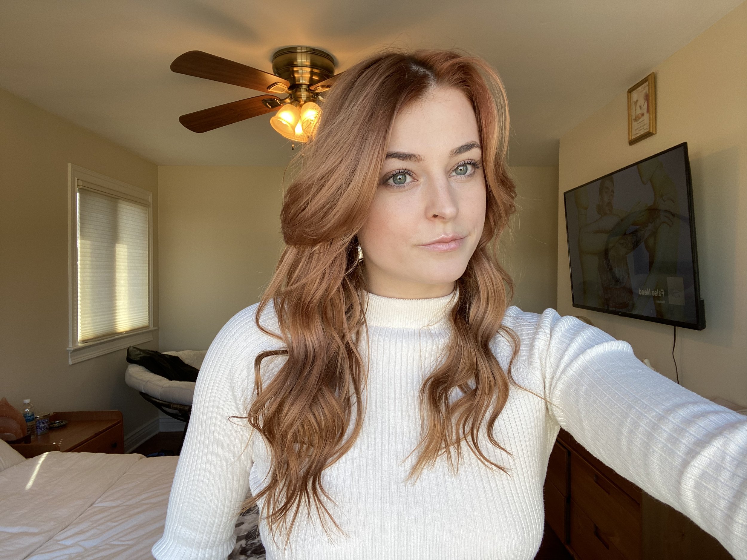 A young woman with reddish-brown wavy hair and bright blue eyes taking a selfie in a bedroom, wearing a white ribbed long-sleeve top. The bedroom has beige walls, a ceiling fan with light, and a mounted flat-screen TV showing a paused image of a person on a yellow background.