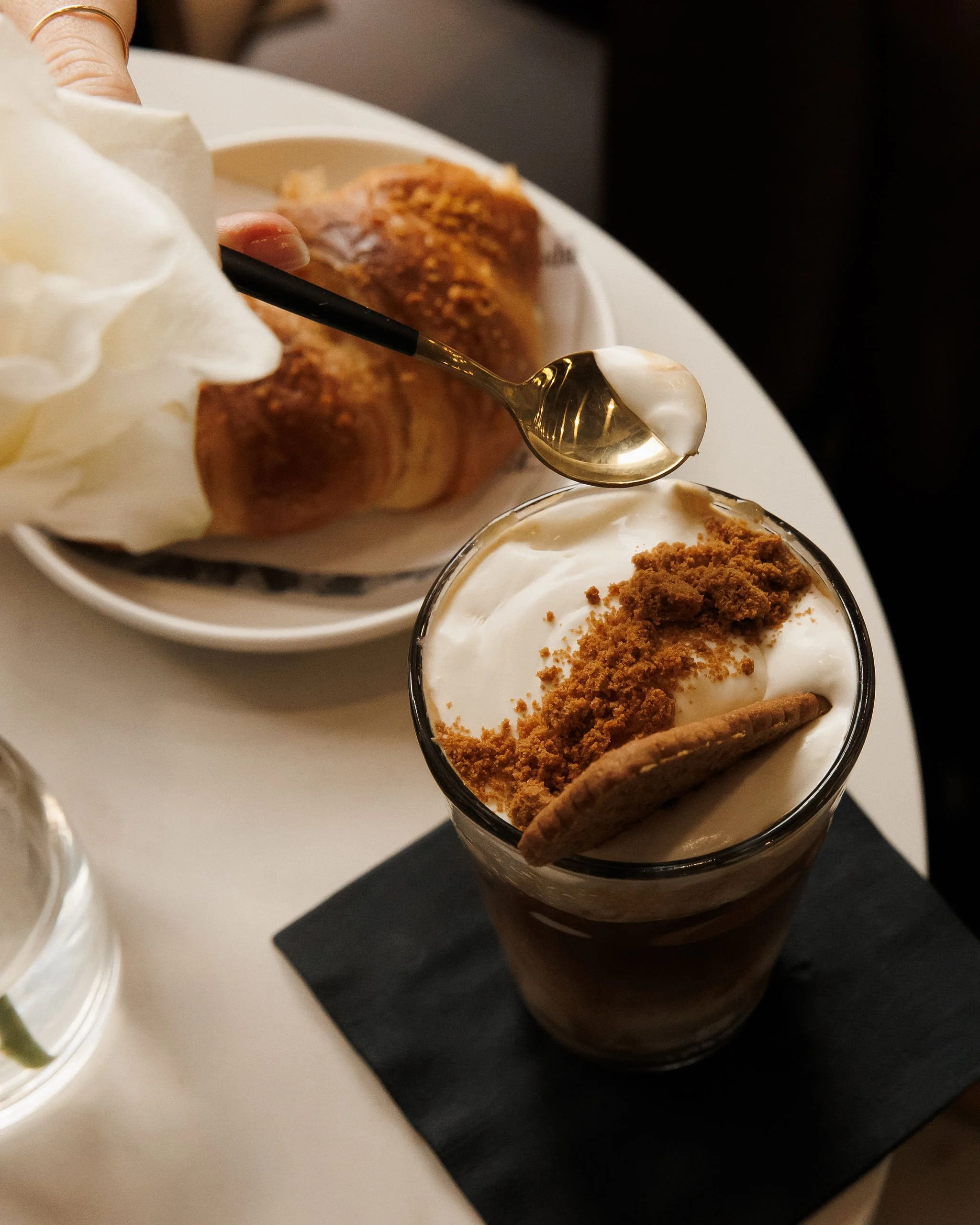 A dessert in a glass topped with whipped cream, cookie crumbs, and a cookie, with a plate of bread rolls and whipped cream in the background.