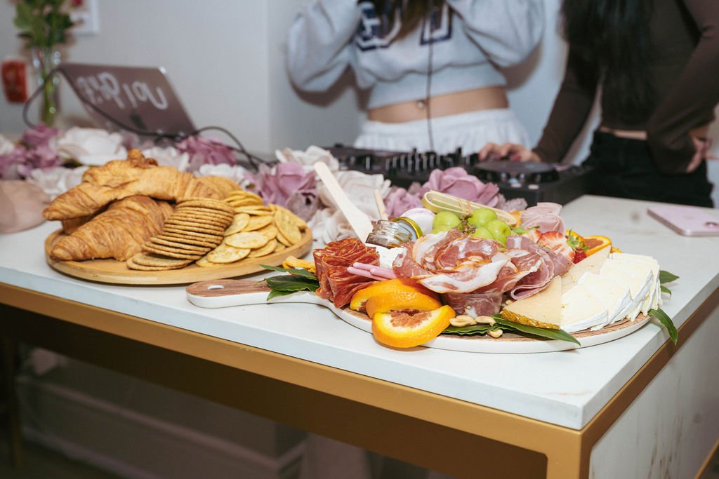 A table with a platter of assorted meats, cheeses, and fruits, alongside a plate of croissants, crackers, and potato chips. In the background, two people are DJing.
