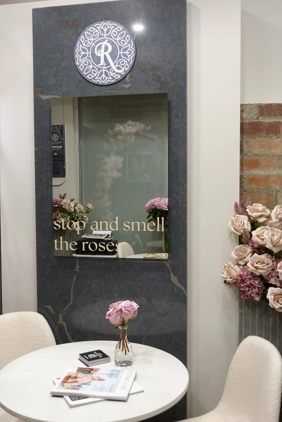 Interior of a flower shop with a round table, a small vase with pink roses, magazines, and a black box on the table. Behind the table is a mirror with gold lettering that says 'stop and smell the roses'. To the right, there is a large flower arrangem
