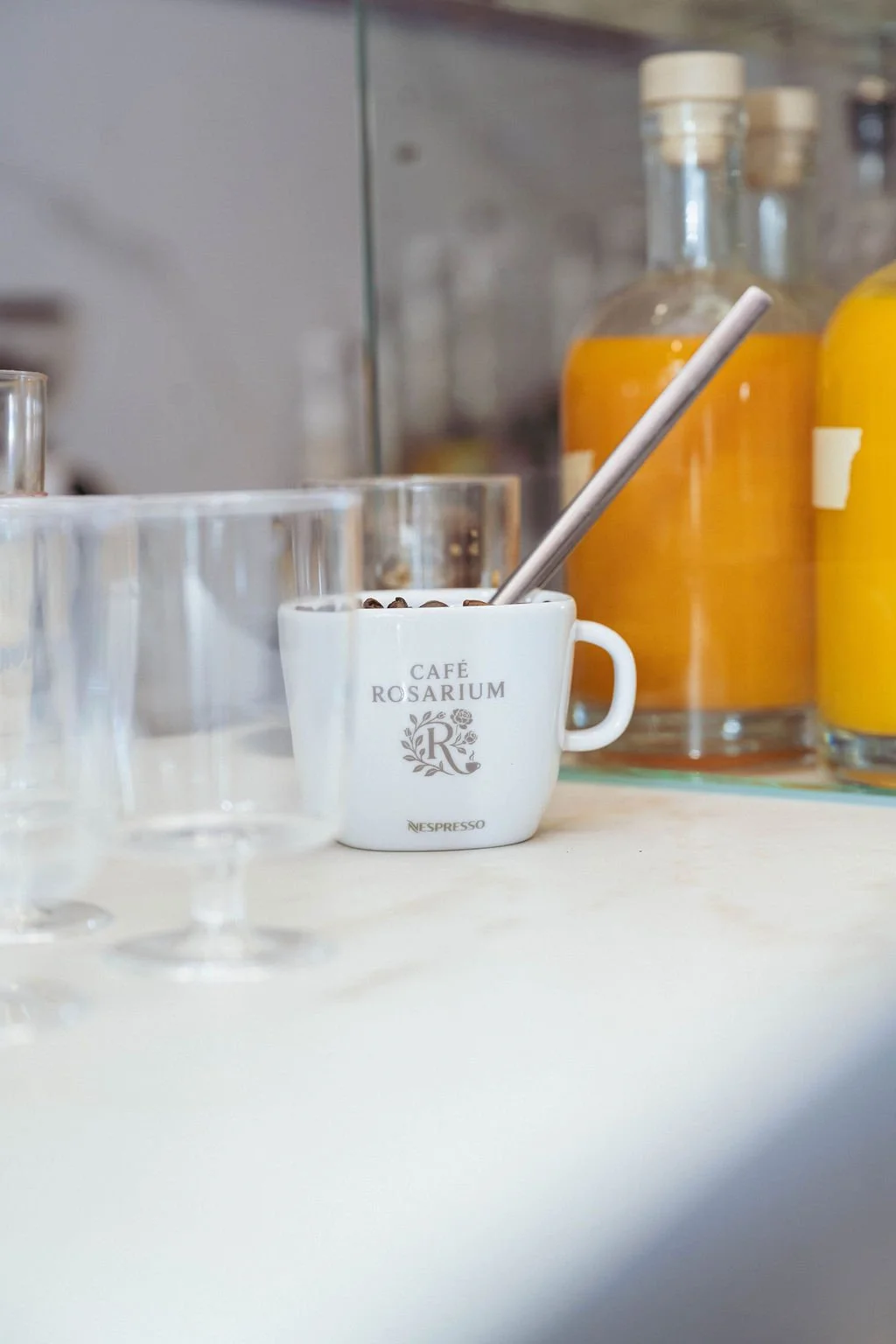 A white mug labeled Café Rosarium with coffee beans and a metal stir stick inside, placed on a light-colored countertop, with glass bottles containing orange juice in the background.