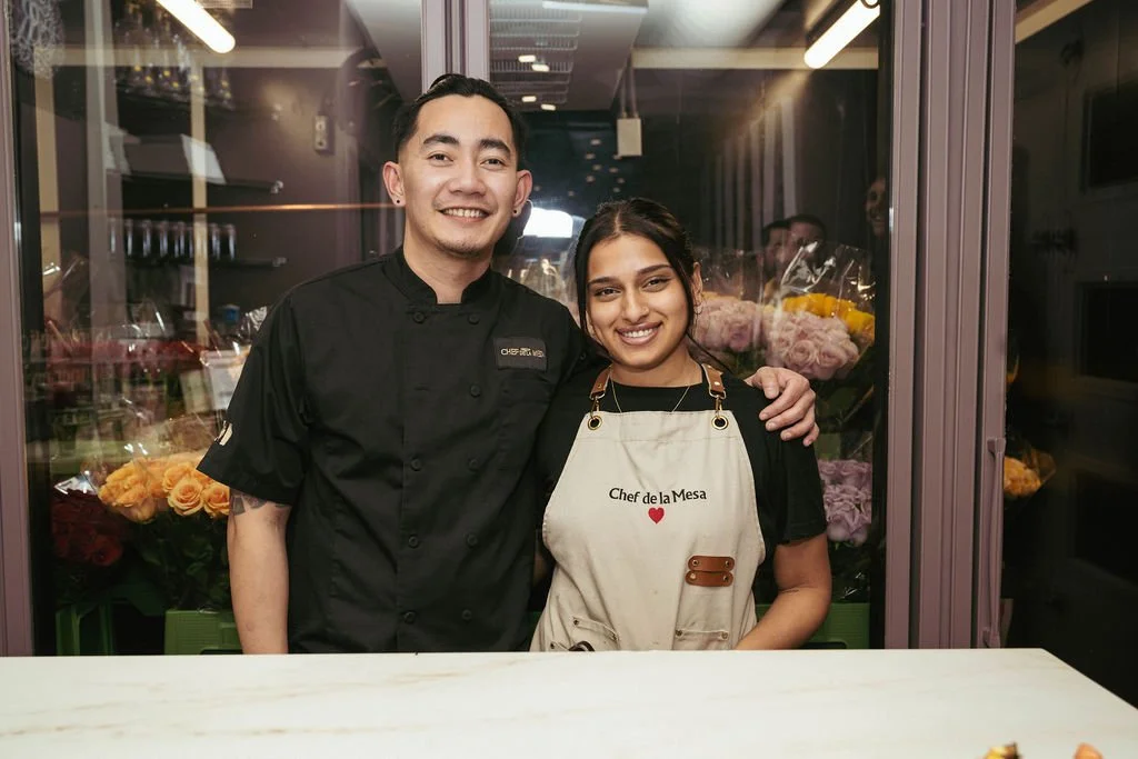 A smiling man in a black chef's coat with a woman in a black shirt and apron that says "Chef de la Mesa" standing together in a kitchen or restaurant setting, with flowers visible through the glass behind them.