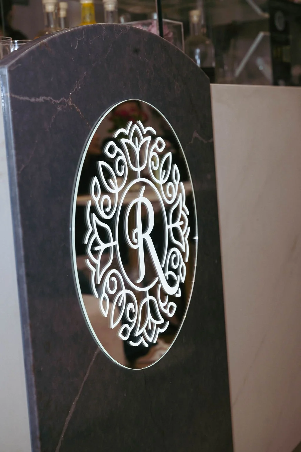 Close-up of a decorative sign with a black marble background and a white illuminated monogram featuring the letter 'R' surrounded by floral patterns.