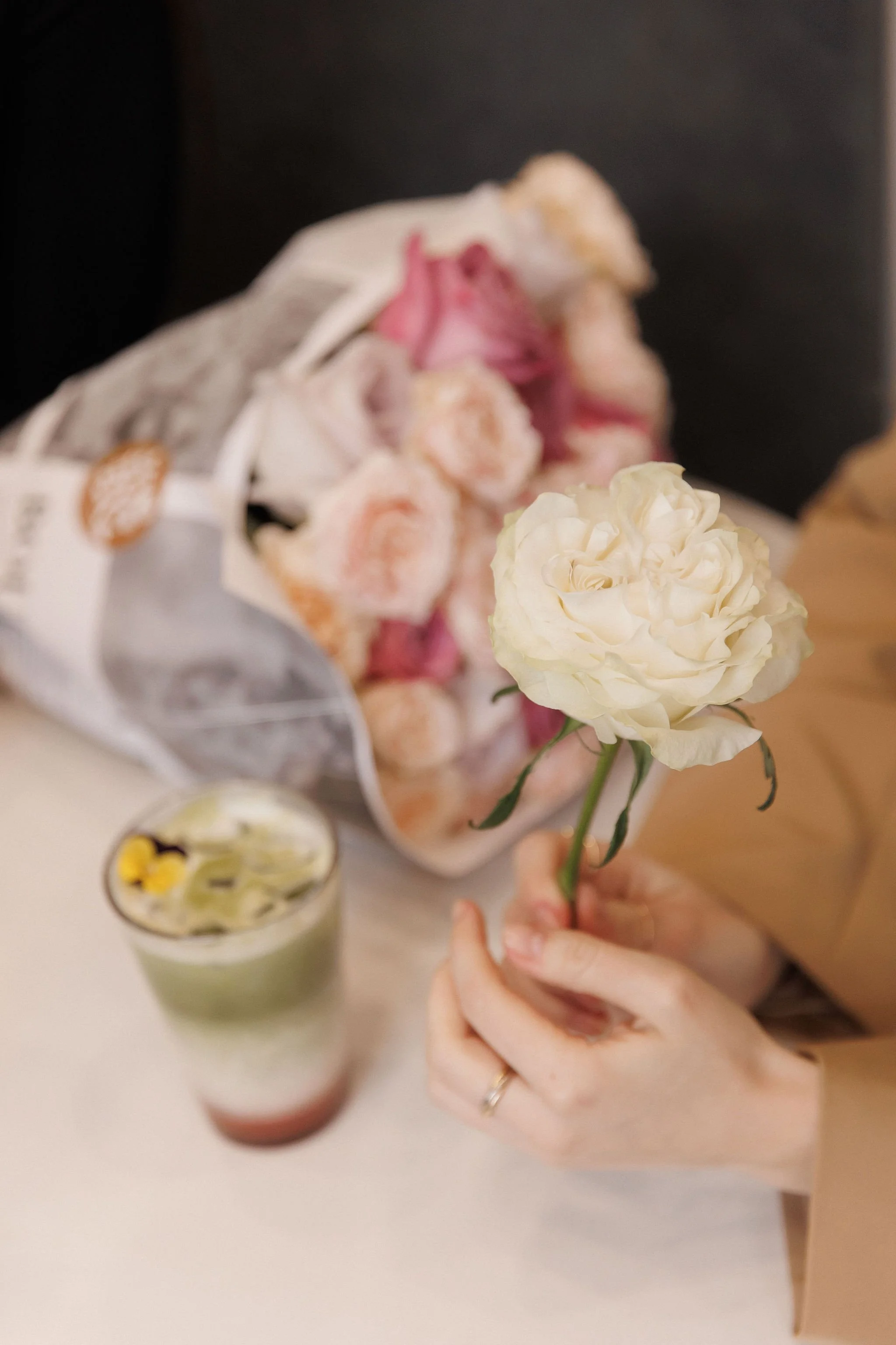 Person holding a white rose with a bouquet of pink roses and a layered drink on a table.