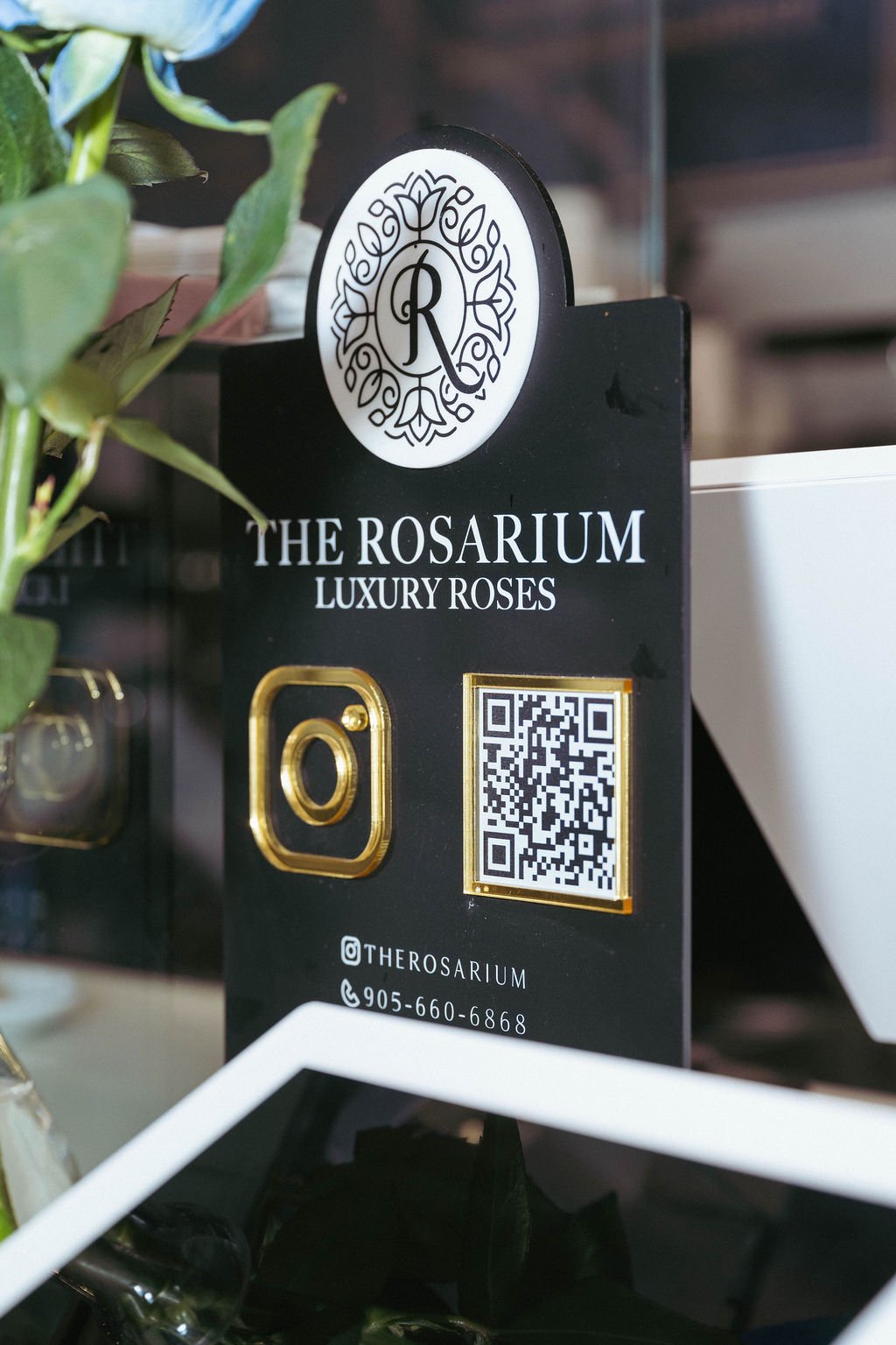 Business card or promotional display for The Rosarium, a luxury roses business, featuring a logo with stylized roses, the name "The Rosarium Luxury Roses," a gold Instagram icon, a QR code, and contact information including Instagram handle and phone