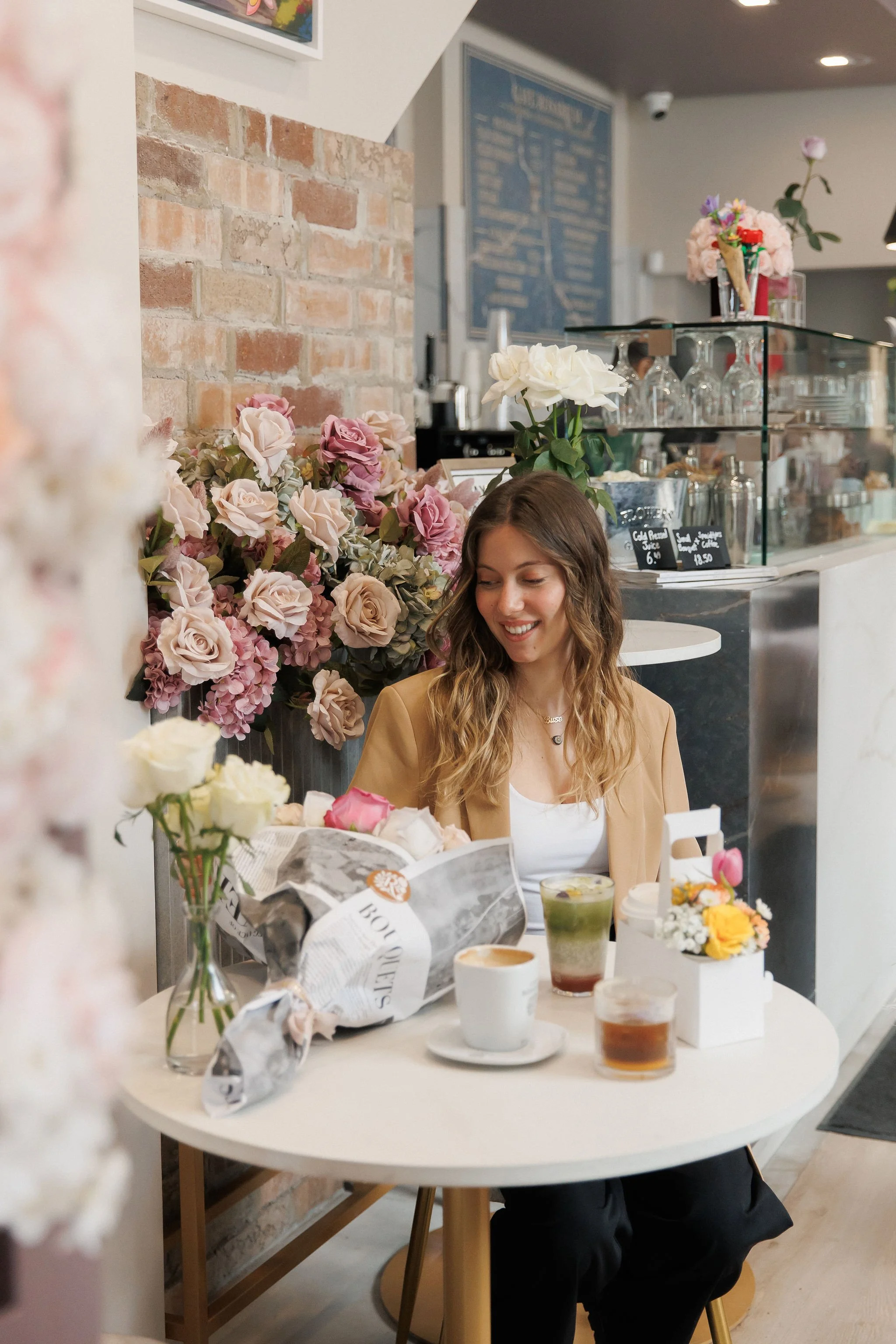 A woman with wavy hair smiling at a table in a cafe adorned with pink and white flowers, various drinks, and pastries, with a brick wall and cafe counter in the background.