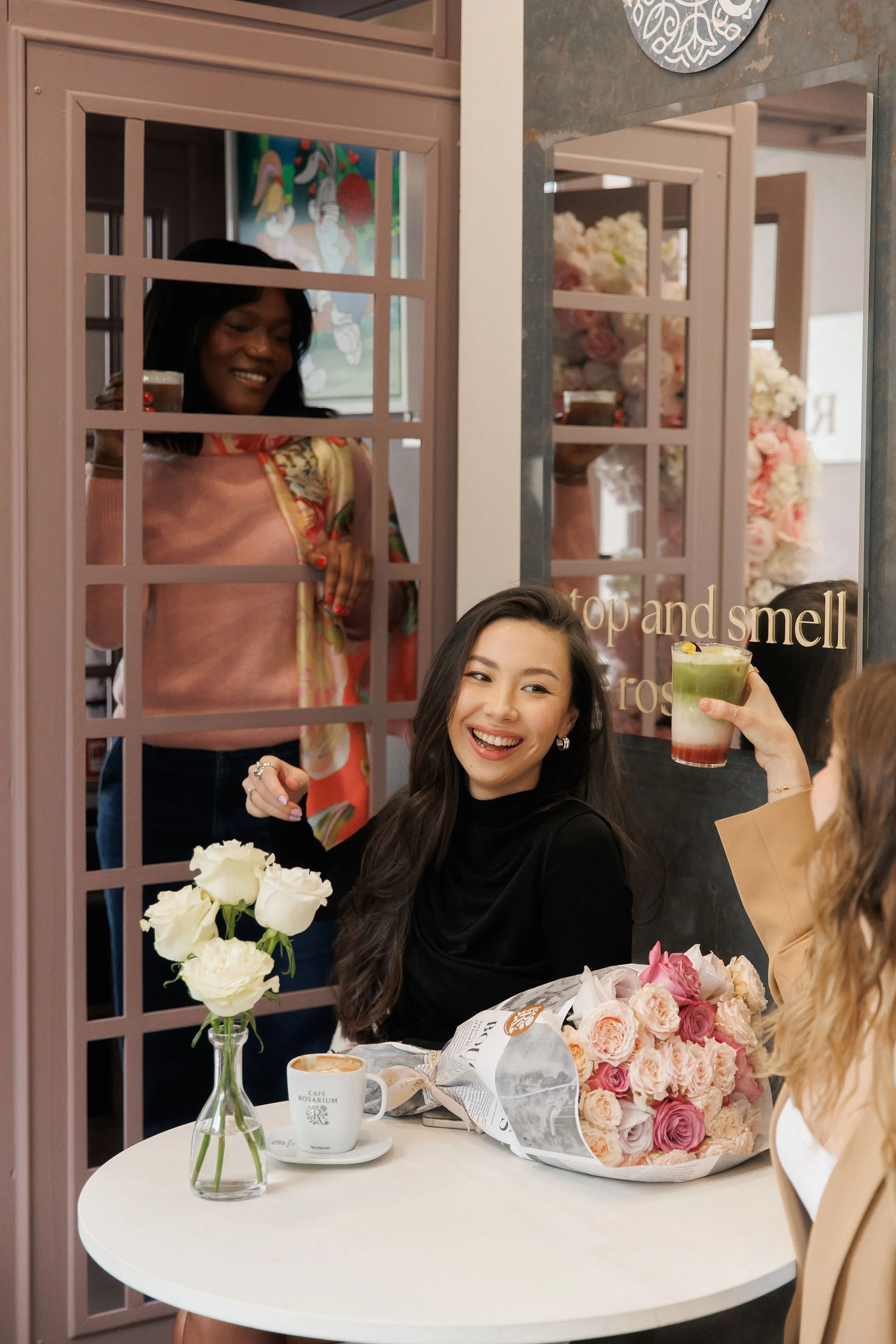 Group of women celebrating with drinks and flowers in a cozy cafe decorated with flowers and pink accents.