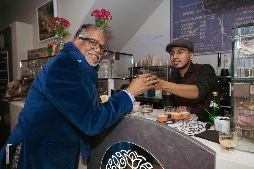 A man with gray hair and glasses wearing a blue velvet jacket and scarf is smiling as he receives a drink from a barista in a black shirt and cap behind a cafe counter. The background includes flowers, glassware, and a menu board.