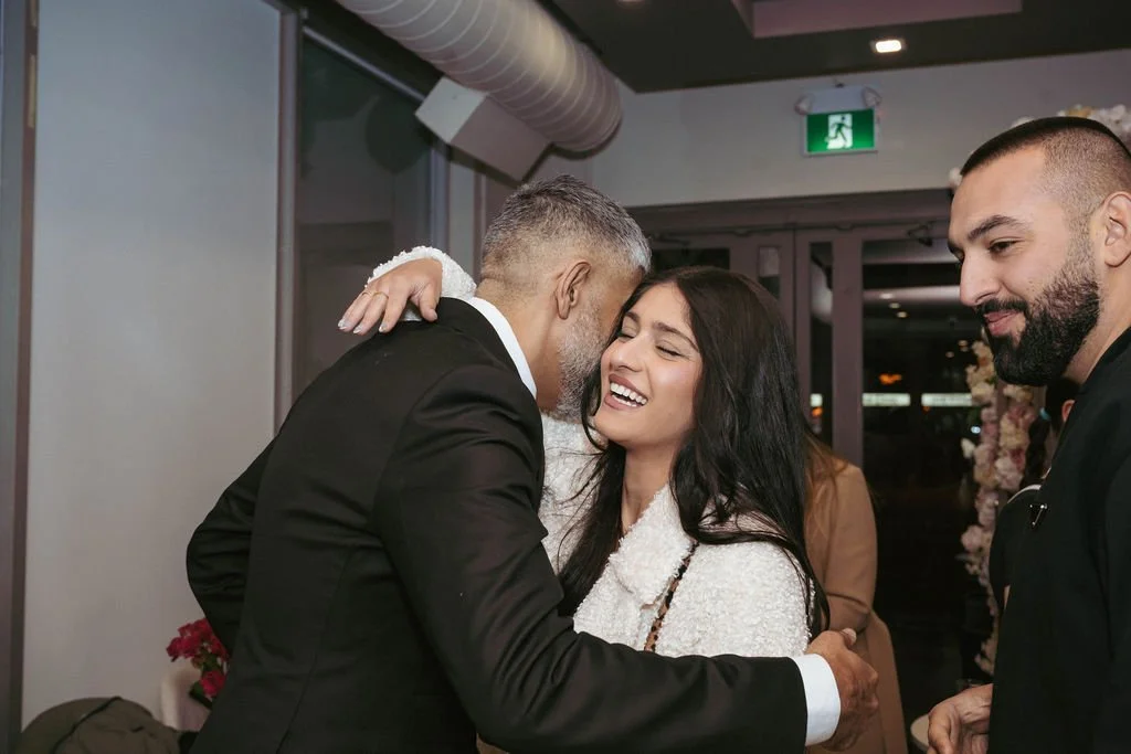 A joyful woman hugging an older man in a suit at a celebration, with a man with a beard standing nearby in a black outfit against a decorated indoor backdrop.