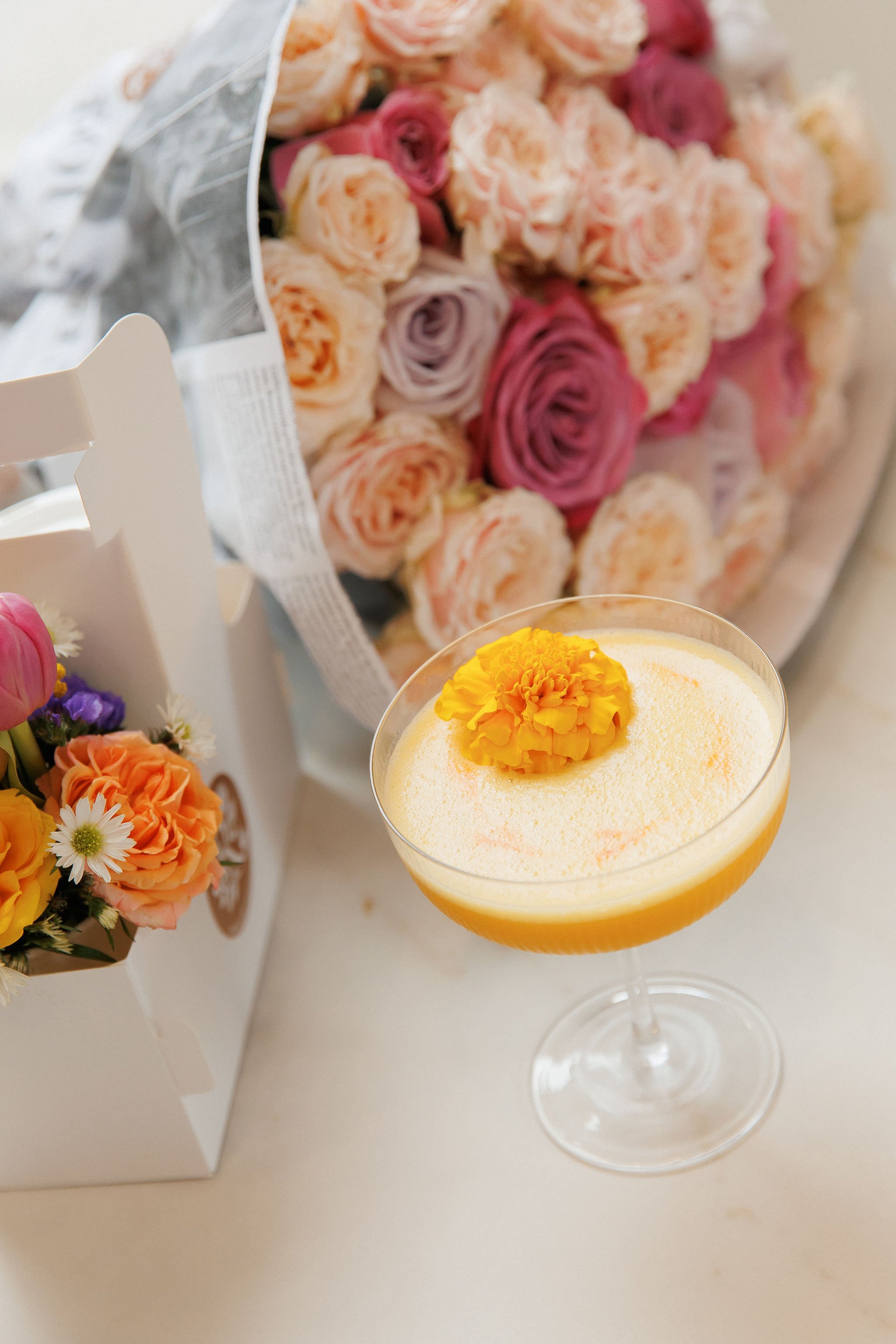 A cocktail in a coupe glass topped with yellow flower petals, with a large pink flower as decoration. In the background, a bouquet of pink, purple, and orange roses and a white box with smaller flowers.