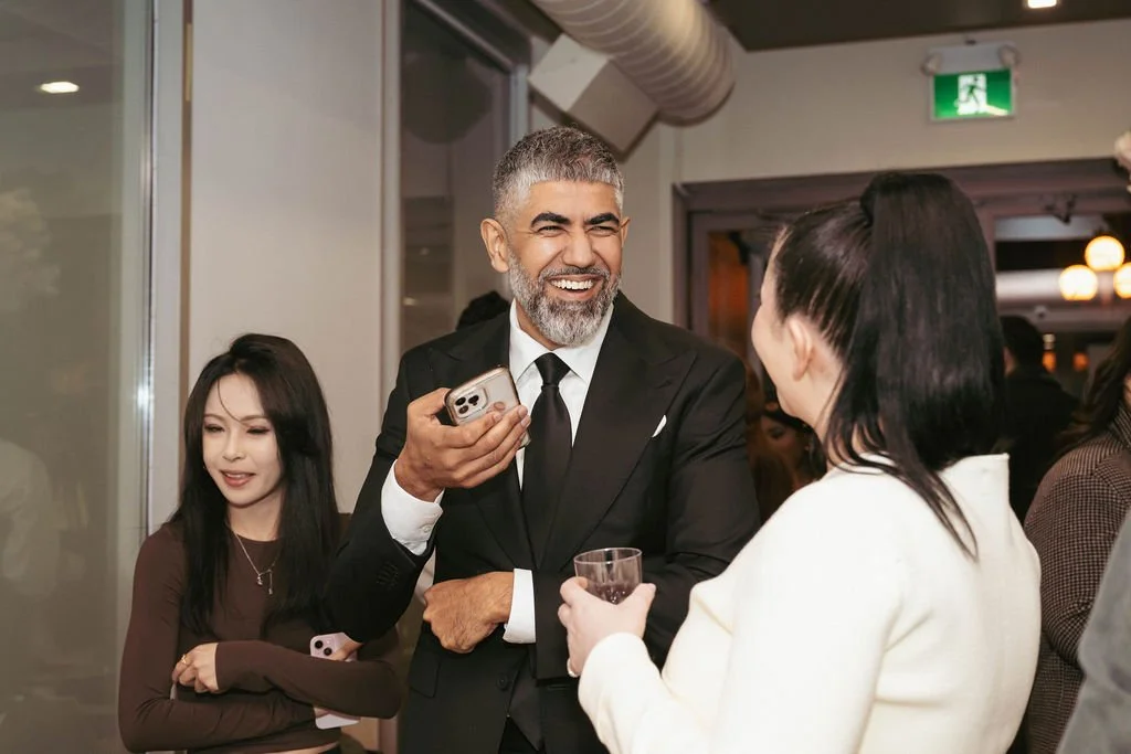 A group of people socializing at a party, with a man in a black suit smiling and holding a phone, and a woman in a white blazer holding a glass, engaged in conversation.