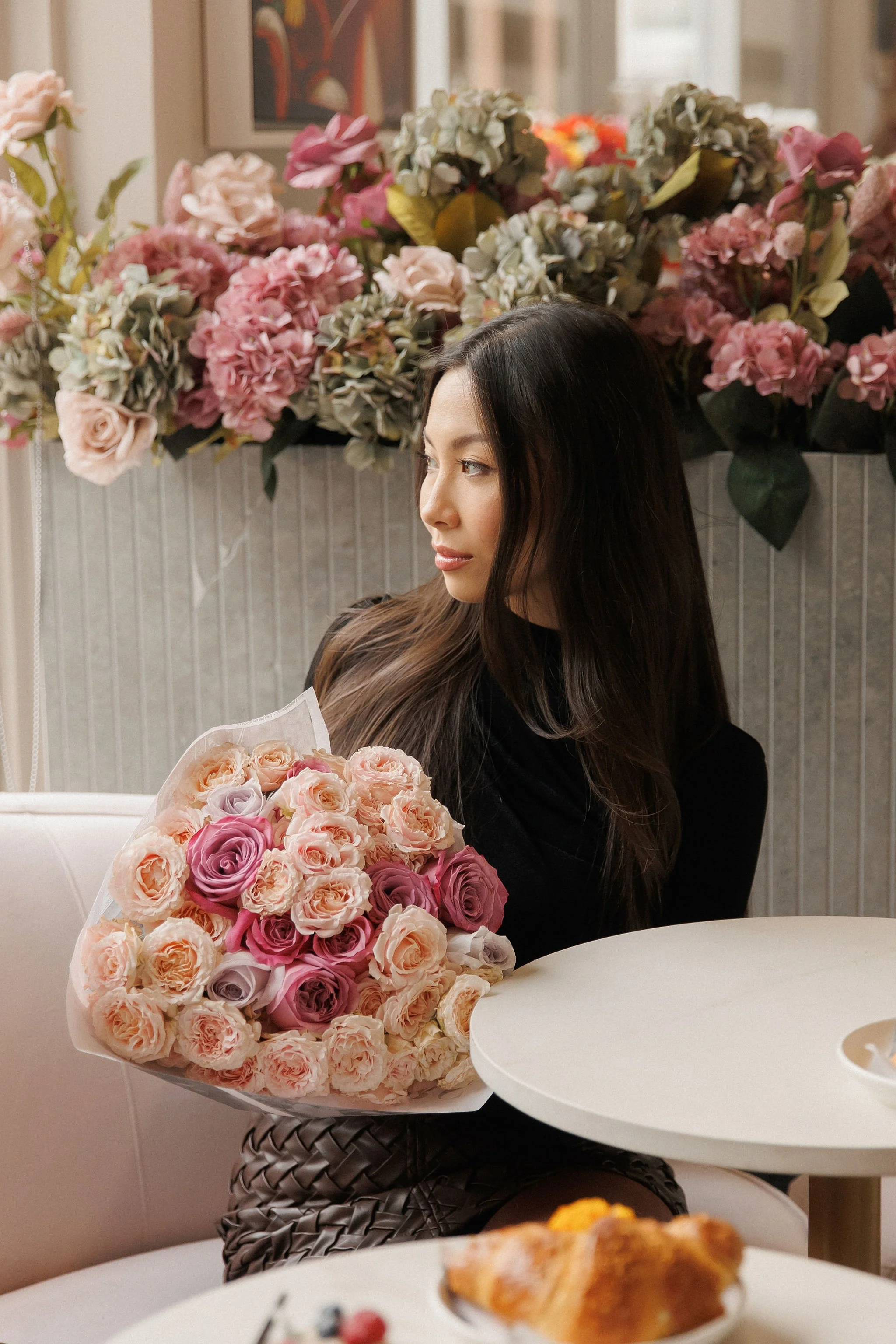 A woman with long dark hair sitting at a table holding a bouquet of pink and purple roses, with a background of pink, white, and green flowers.