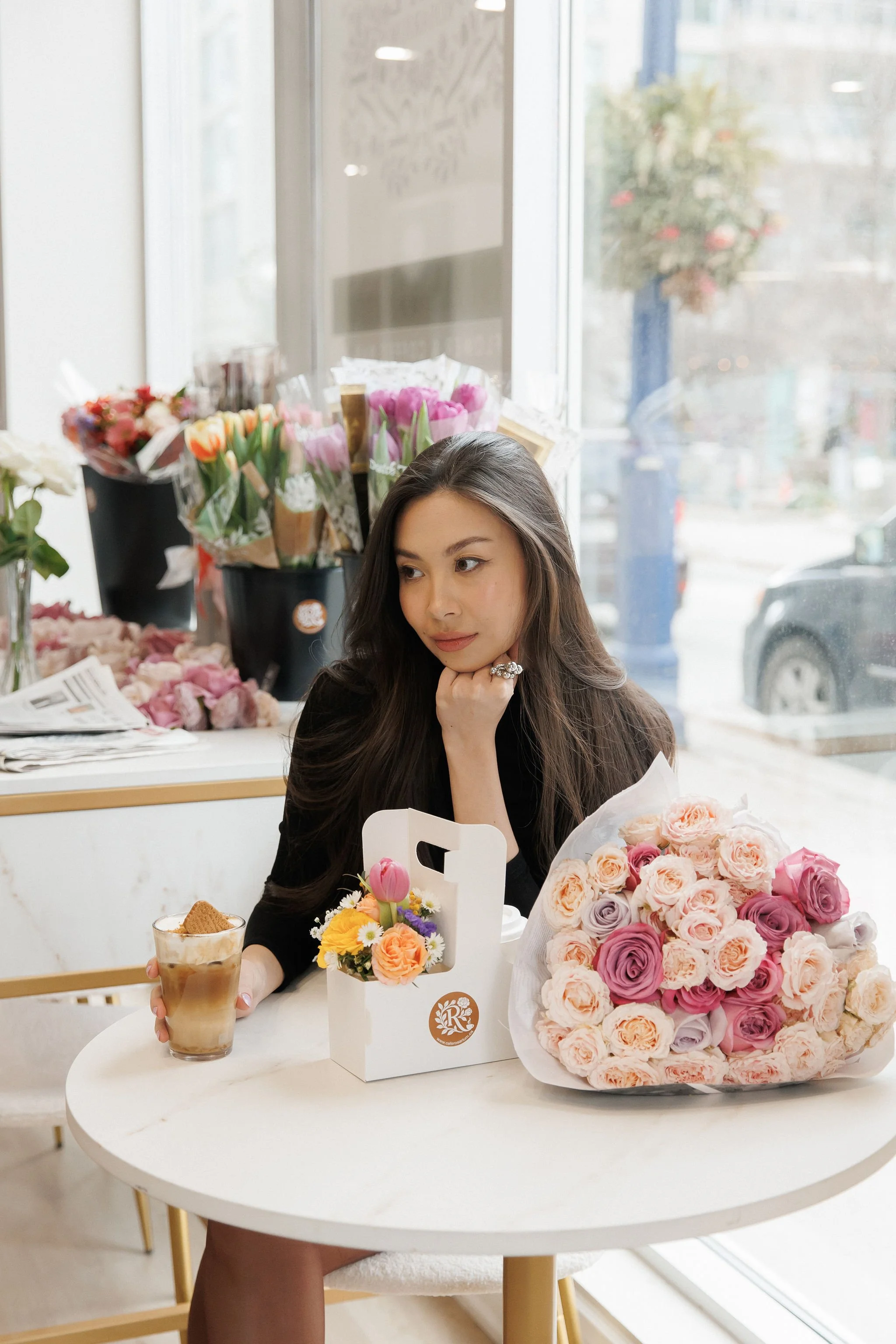 A young woman sits at a white table in a flower shop, surrounded by bouquets of pink and peach roses. She rests her chin on her hand, looking thoughtfully to the side. On the table, there is a large bouquet of roses, a small flower arrangement, a gla