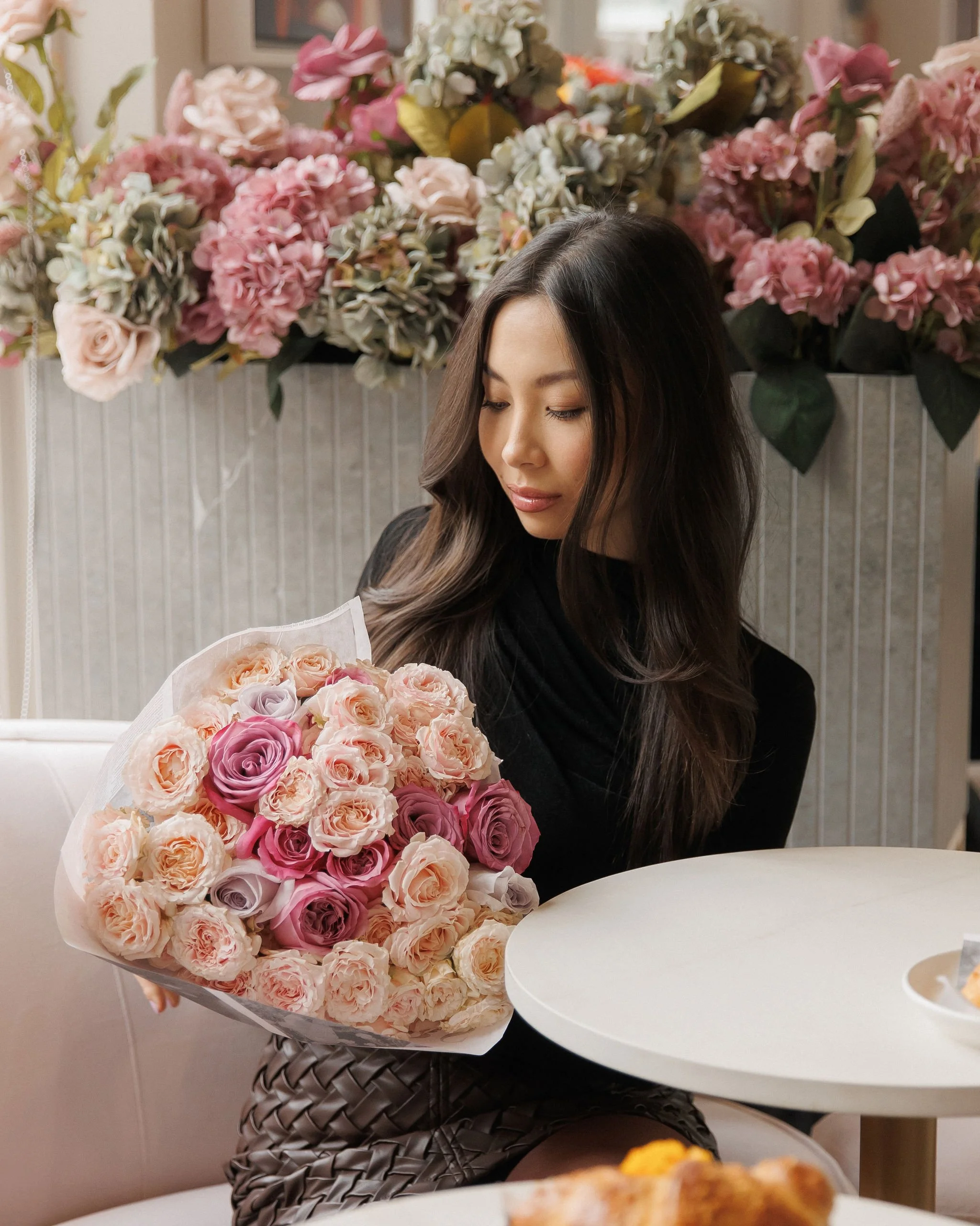 A woman with long dark hair holding a bouquet of pink and cream roses, sitting in front of a wall decorated with hydrangea flowers in pink, white, and green, at a table in a cozy indoor setting.