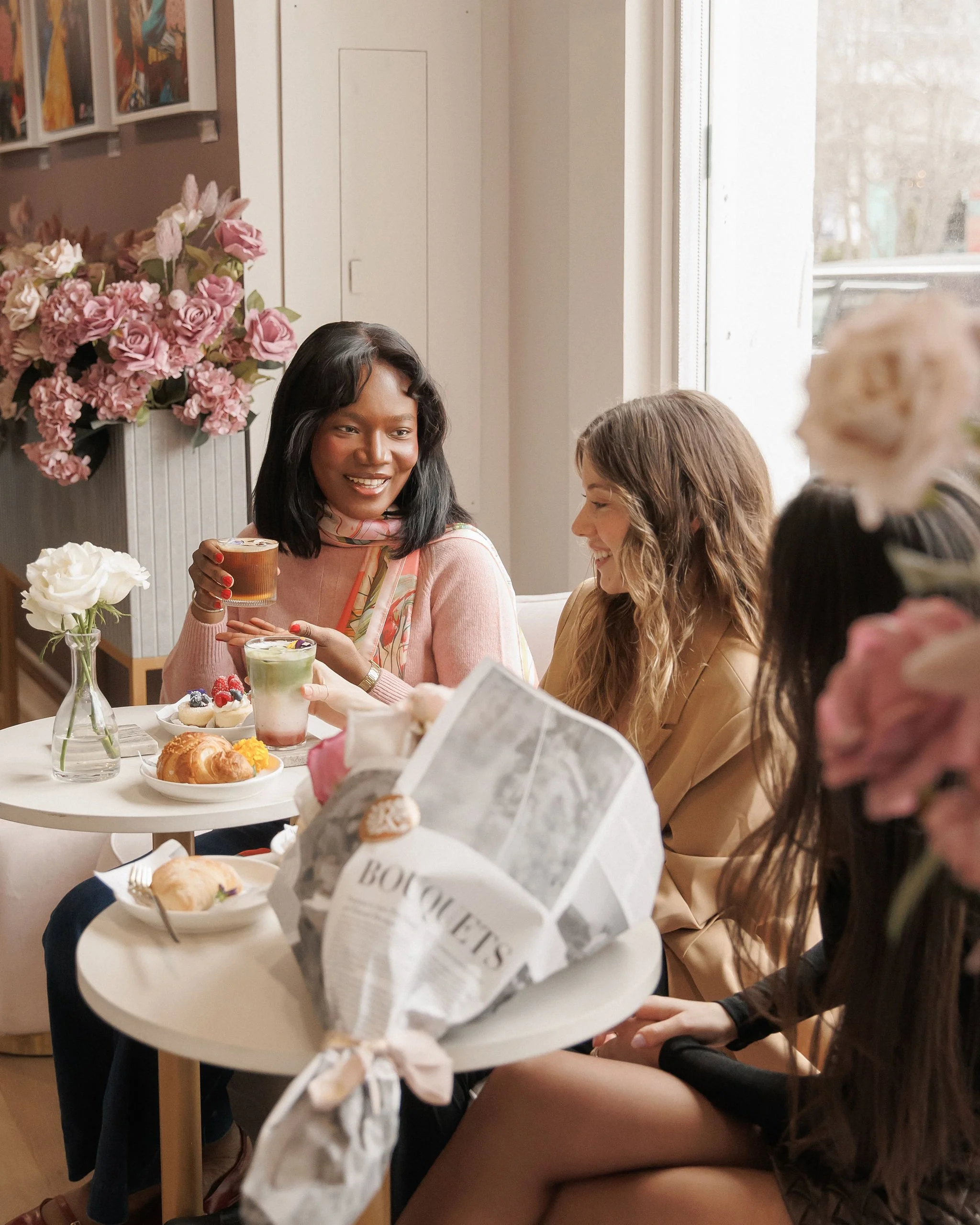 Three women enjoying a tea party with pastries, flowers, and colorful drinks in a cozy cafe.