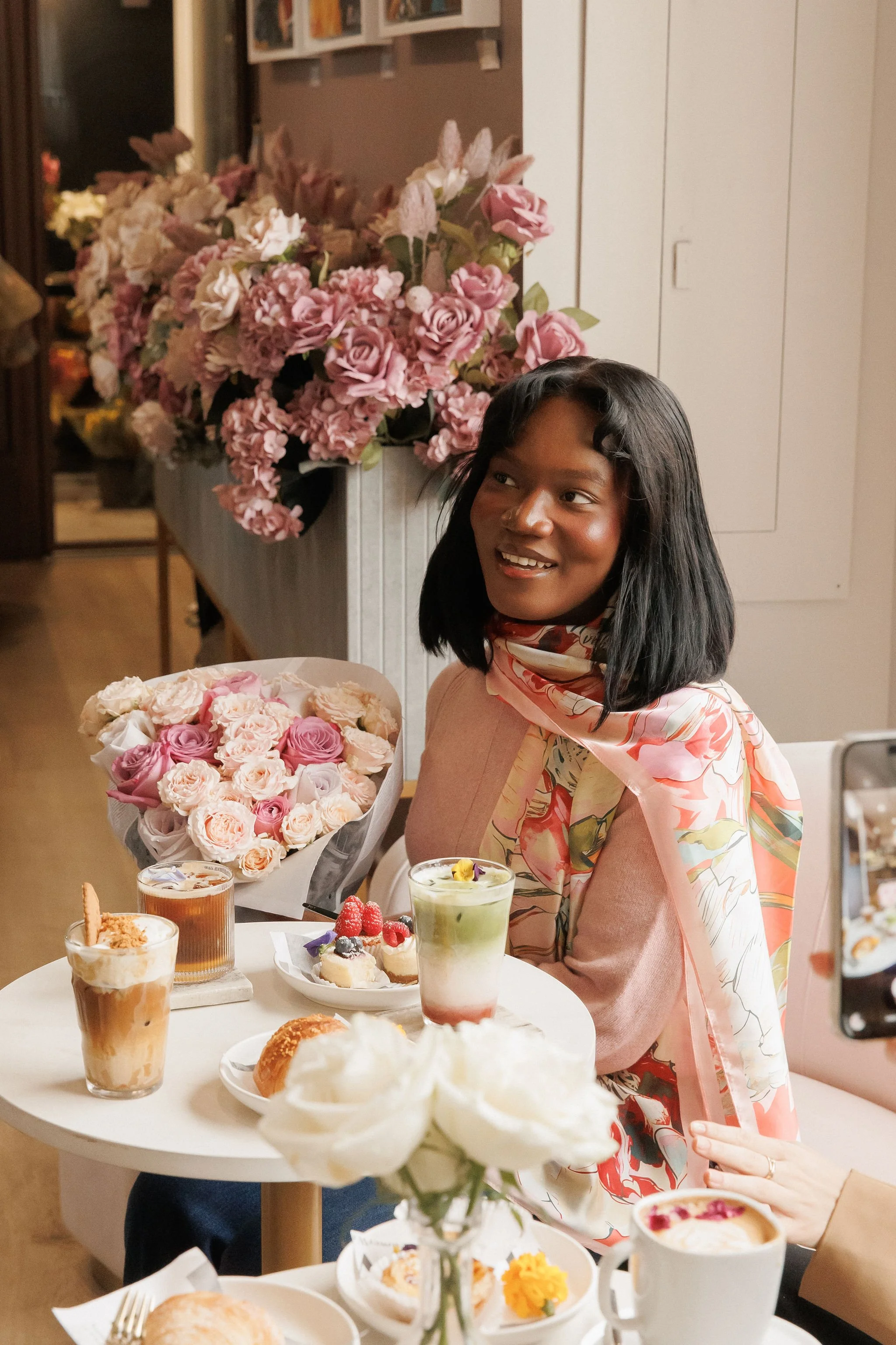 A woman sitting at a table with an assortment of desserts and flowers, smiling, in a cozy indoor setting decorated with pink flowers.
