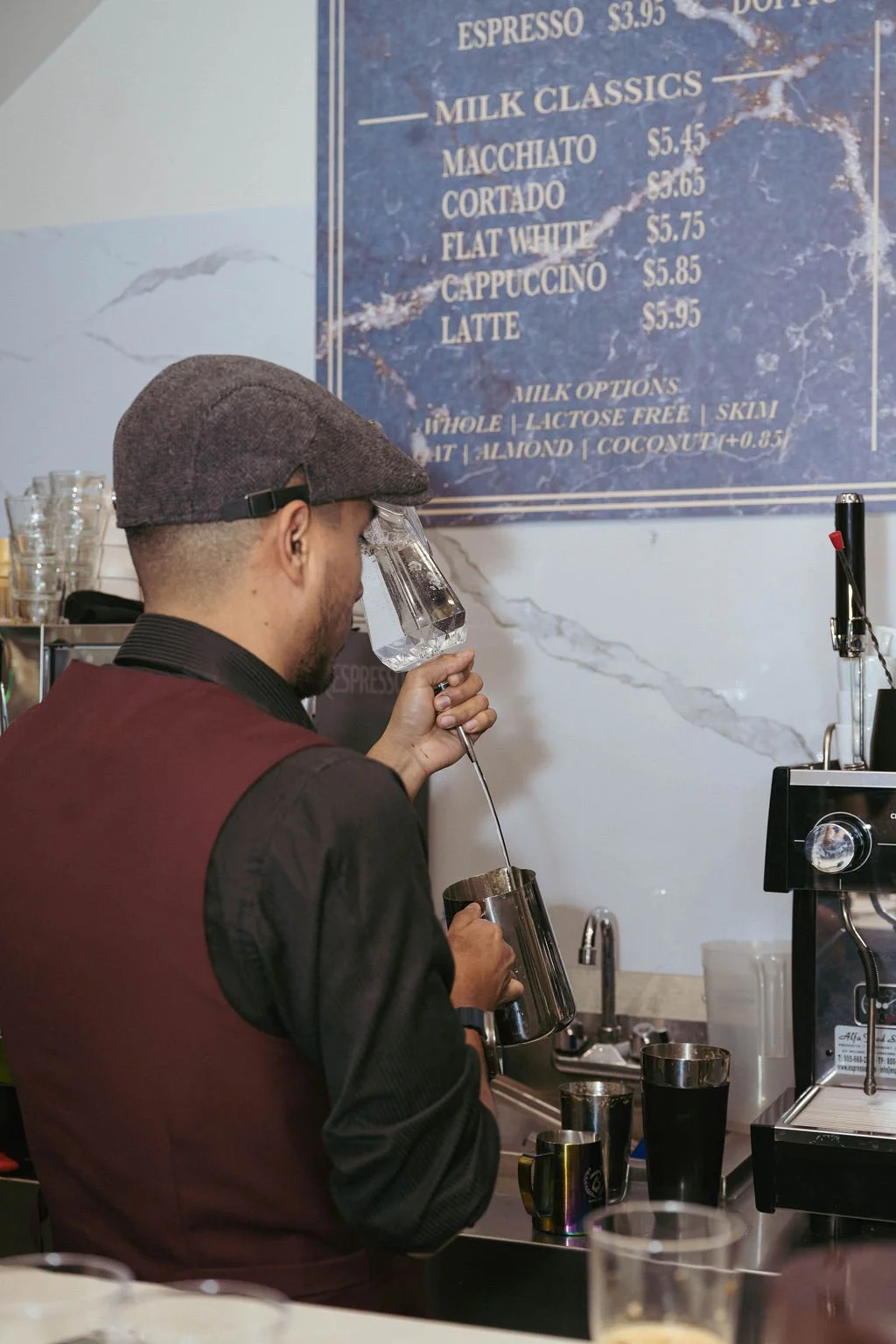 Barista making a drink with a milk frothing pitcher in a coffee shop, with a blue menu board displaying drink options in the background.