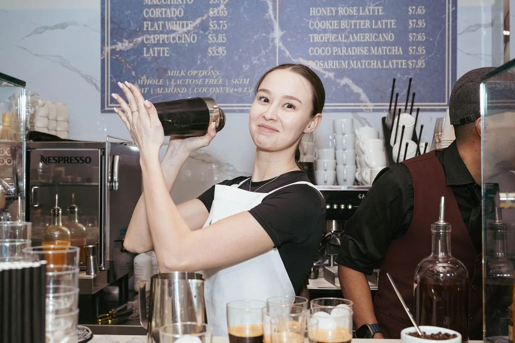 A smiling barista holding a cocktail shaker in a coffee shop with a menu board in the background.
