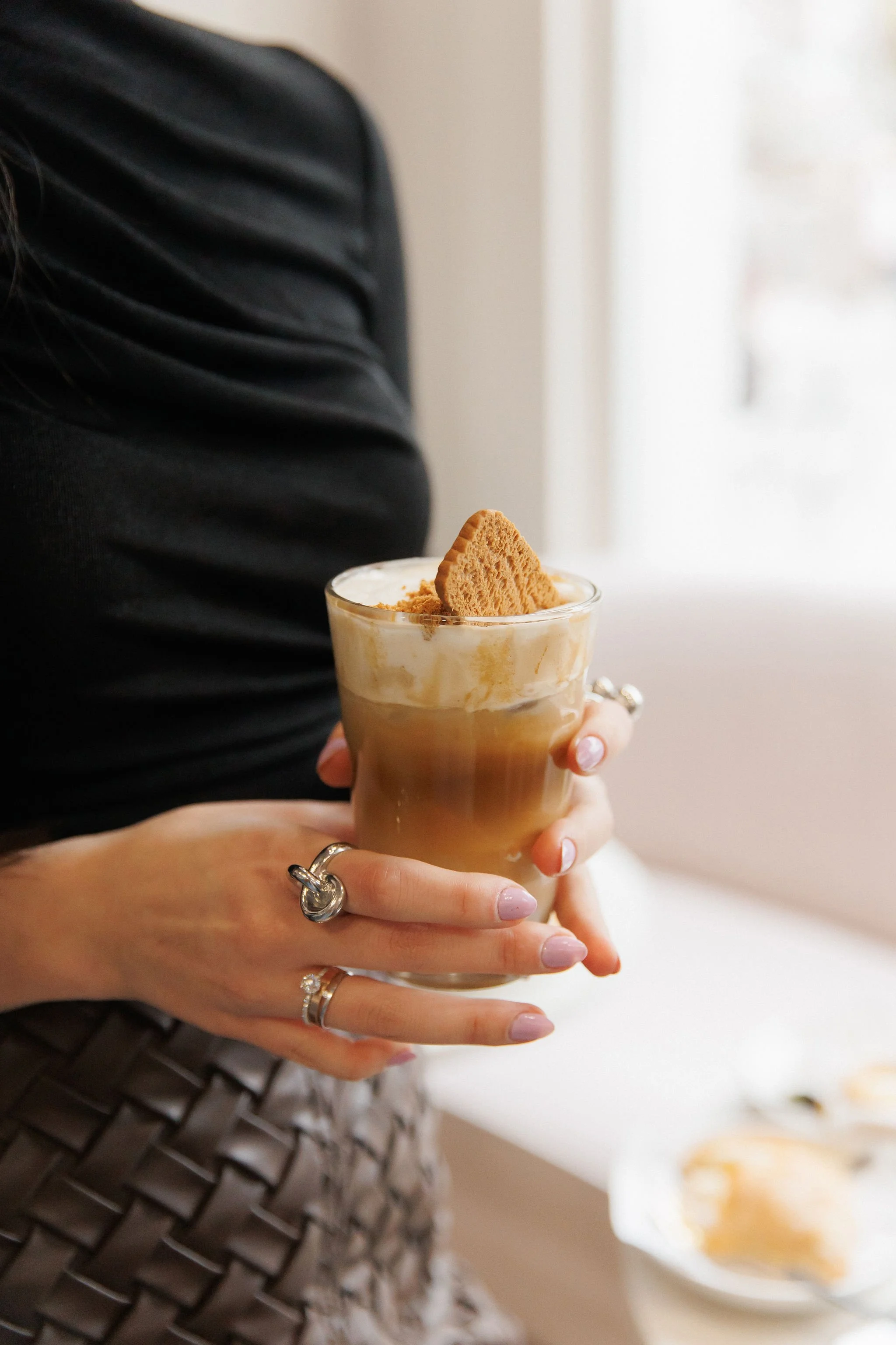 Person holding a glass of iced coffee topped with whipped cream and a biscuit, with a blurred plate of desserts in the background.