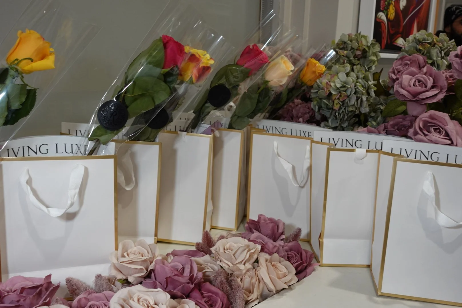 Flowers arranged with gift bags on a table, featuring roses in pink, yellow, cream, and purple, with 'LIVING LUXE' branding visible on bags.