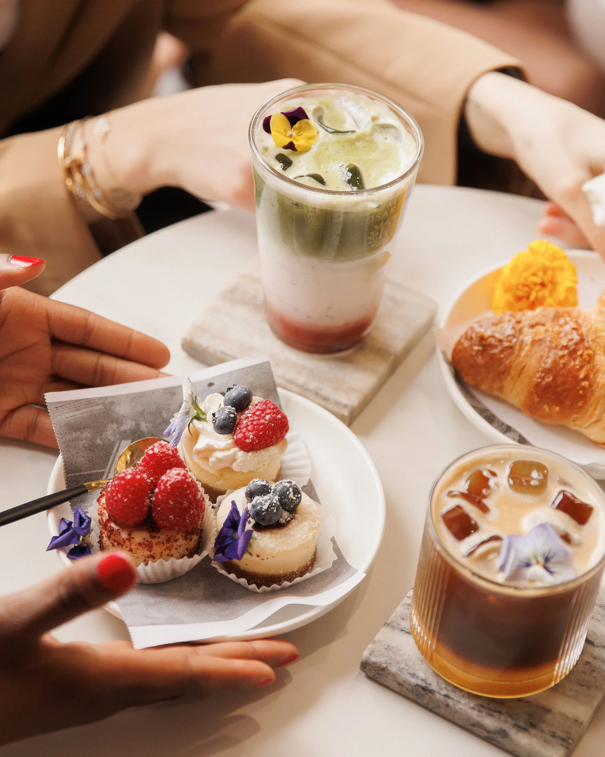 Assorted desserts and beverages on a table, including cupcakes with berries and flowers, muffins, and iced coffee drinks.