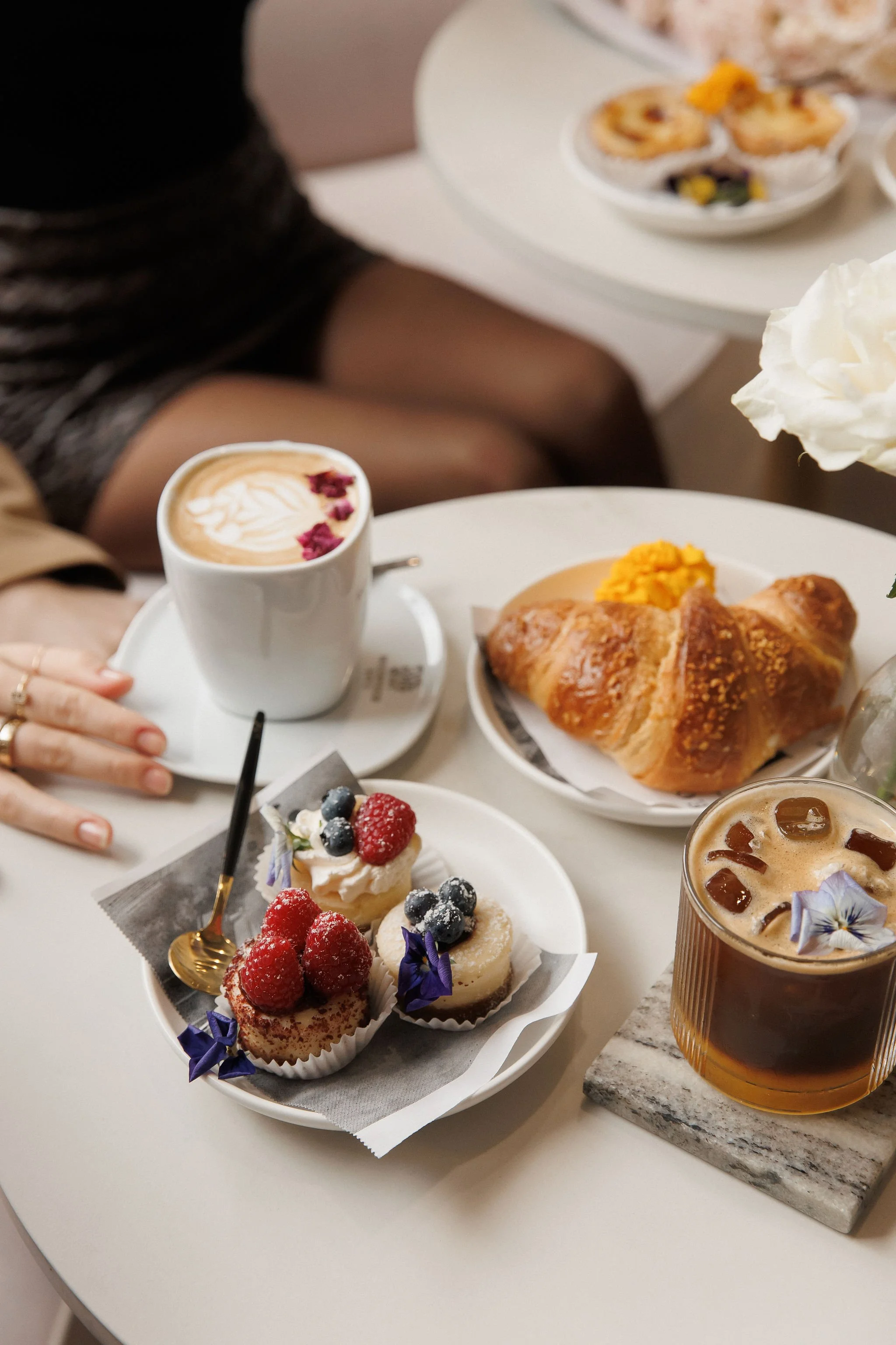 A table with two cups of coffee, a plate with a croissant, a plate with assorted mini desserts topped with berries, and a glass of iced coffee with flowers.