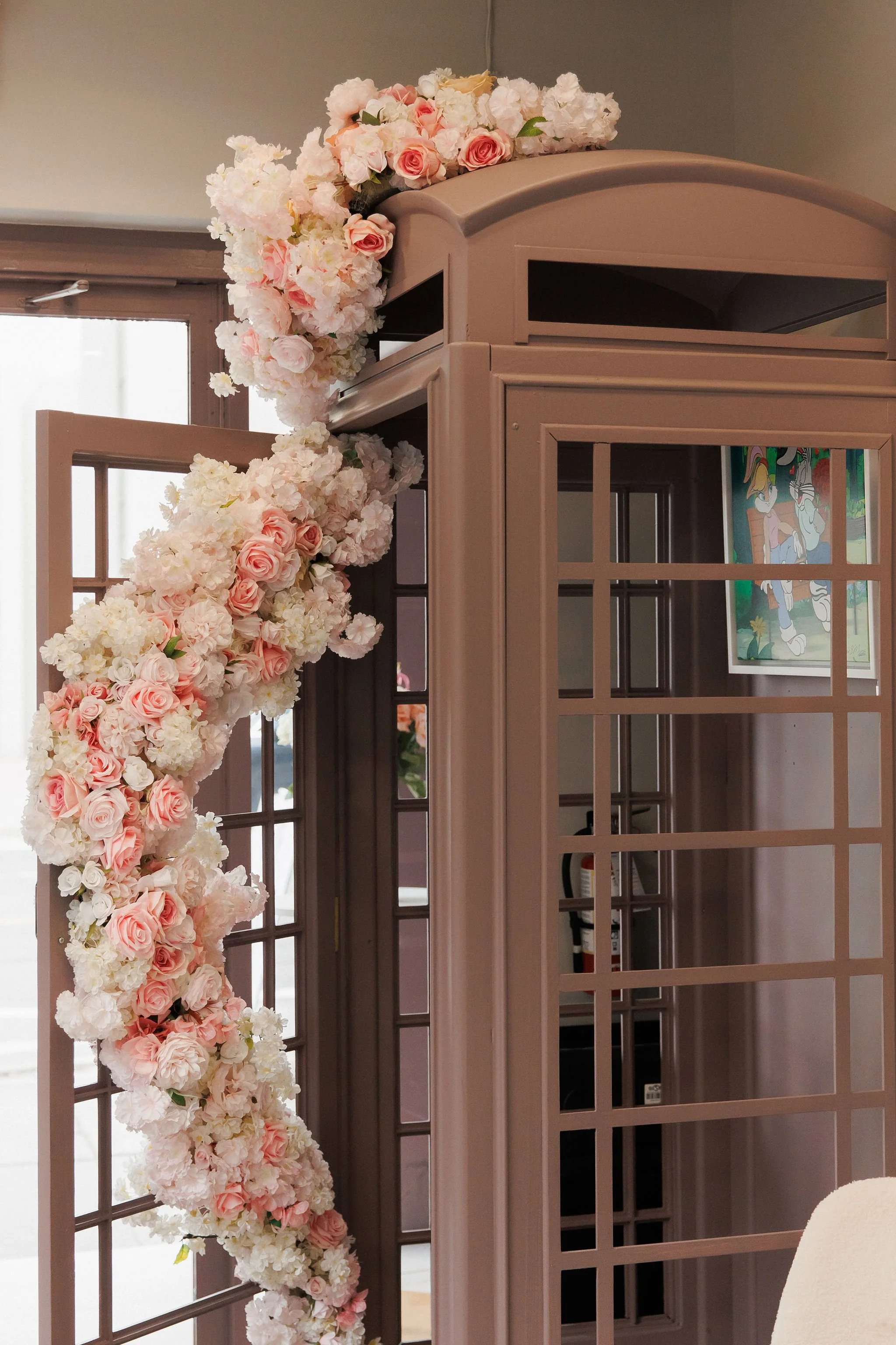 Pink and white wedding arch decorated with an abundance of pink and white flowers, including roses and hydrangeas, inside a room with a door and a picture on the wall.