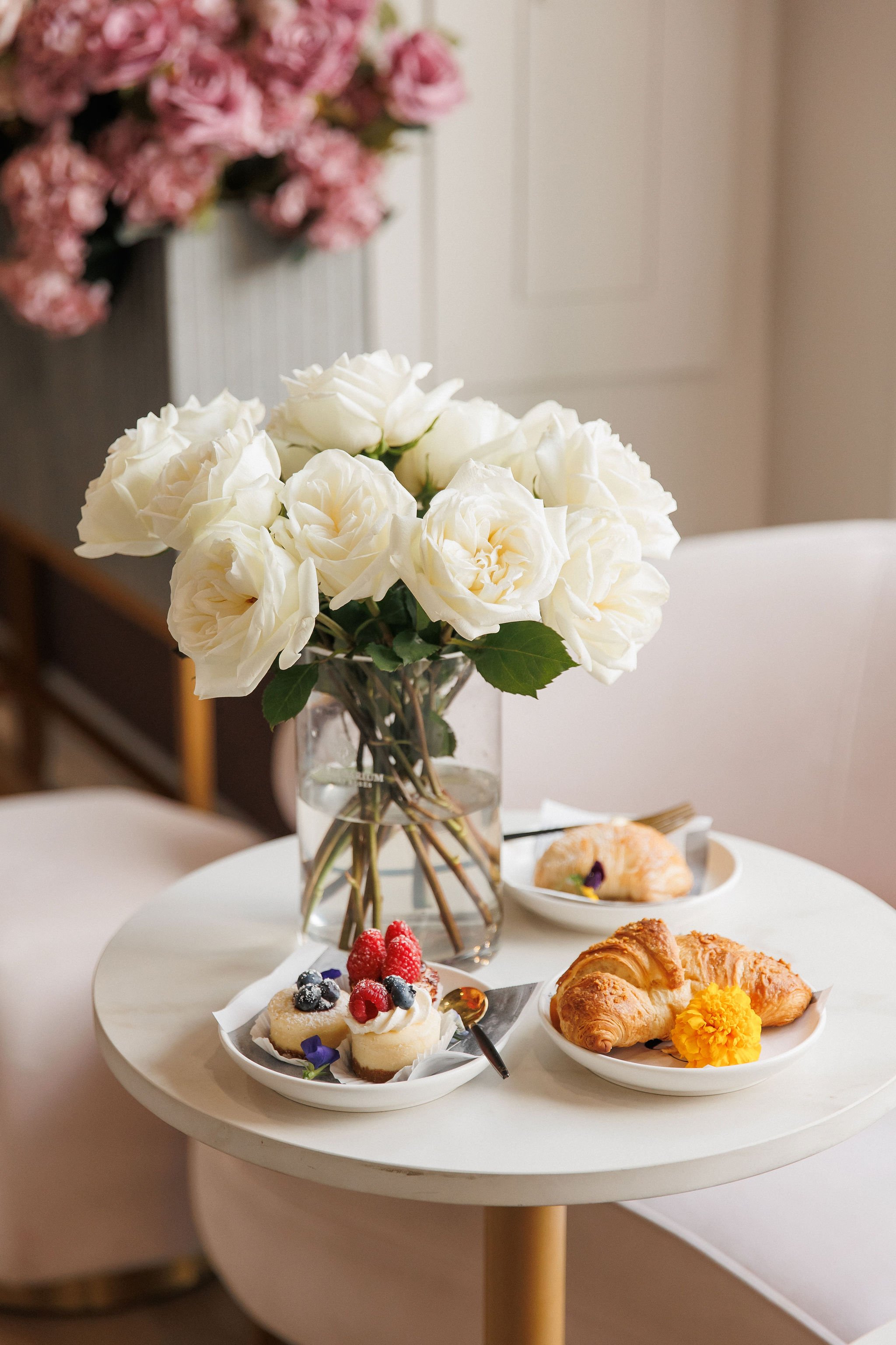 A small round white table with a large glass vase of white roses in the center, surrounded by plates of pastries including croissants and fruit-topped cheesecake, set in a room with pink flowers in the background.