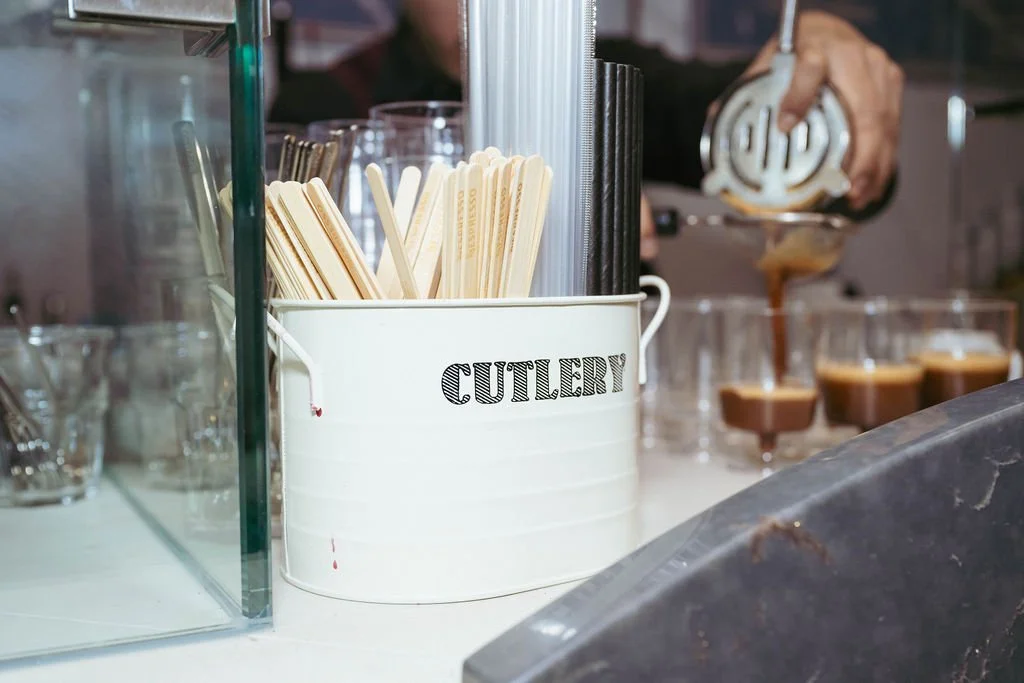 A white container labeled 'cutlery' holds disposable chopsticks and utensils on a counter, with dessert glasses filled with a chocolate dessert in the background.