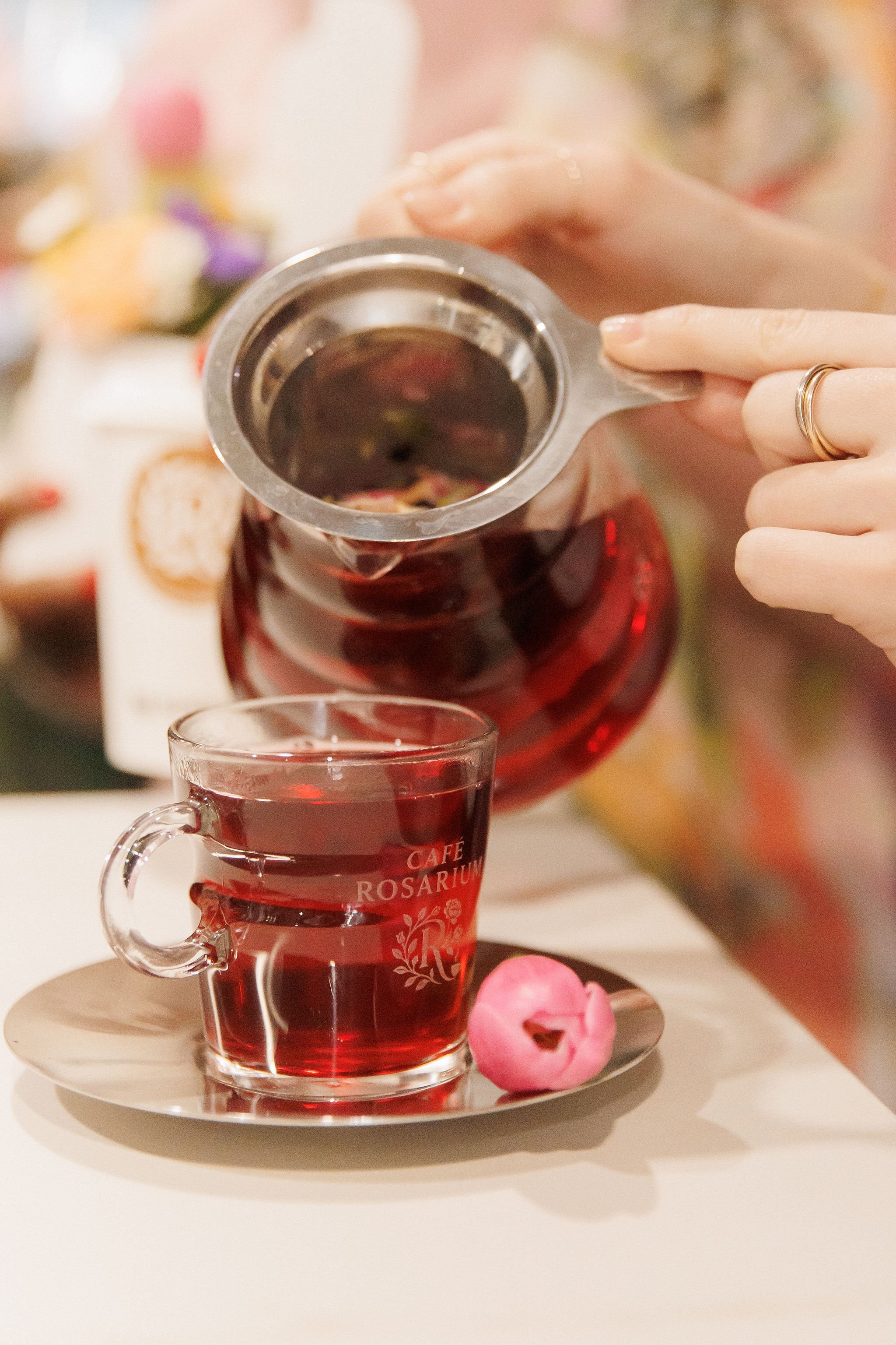 A person pouring tea from a glass teapot into a clear glass cup with a pink flower on a saucer.