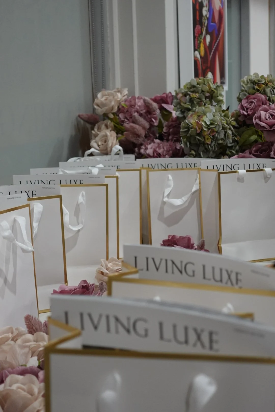 Several white gift bags with the words "Living Luxe" printed on them, surrounded by pink and beige flowers, in an indoor setting.