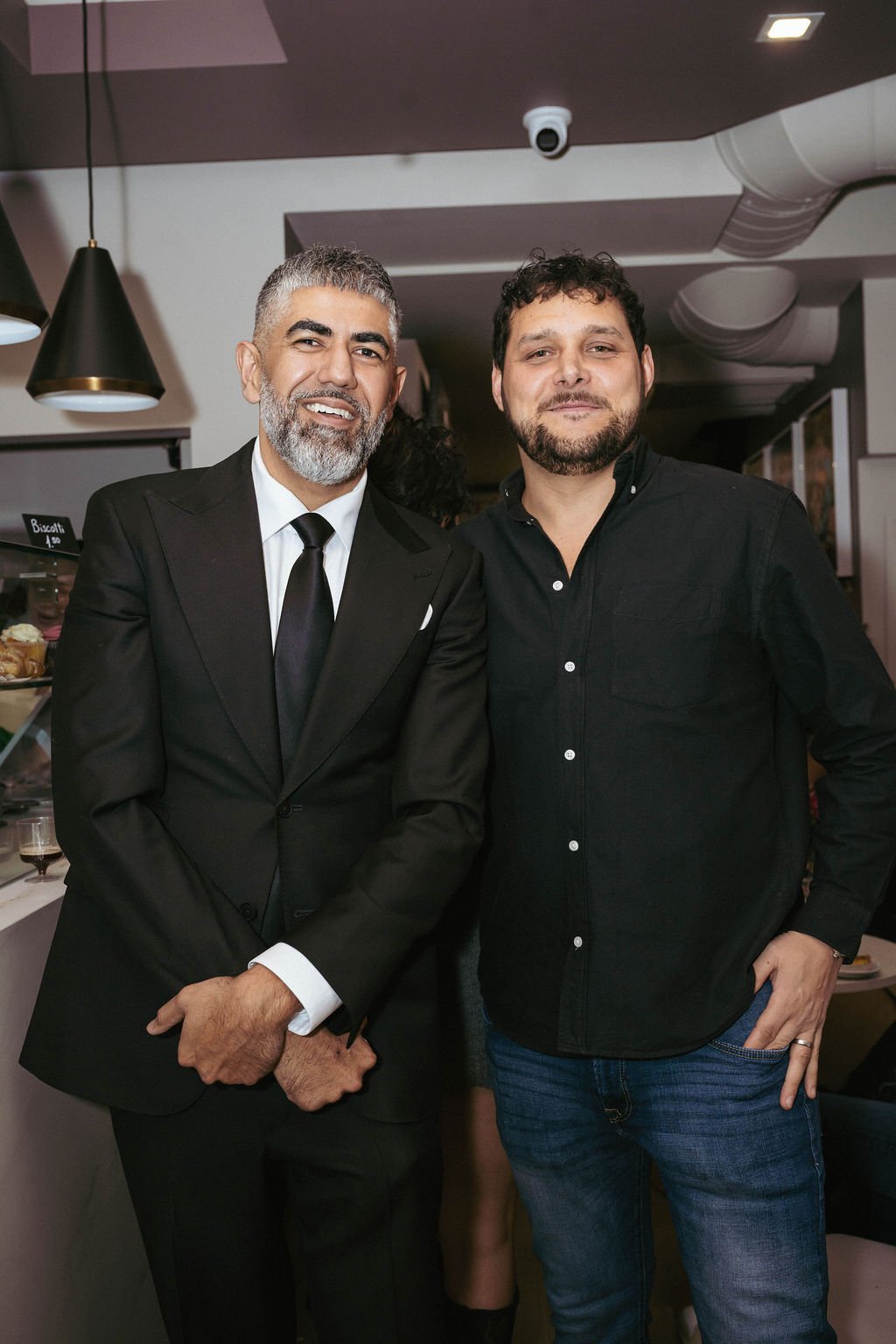 Two men posing for a photo indoors, one in a black suit with a tie and the other in a black shirt and jeans, smiling.