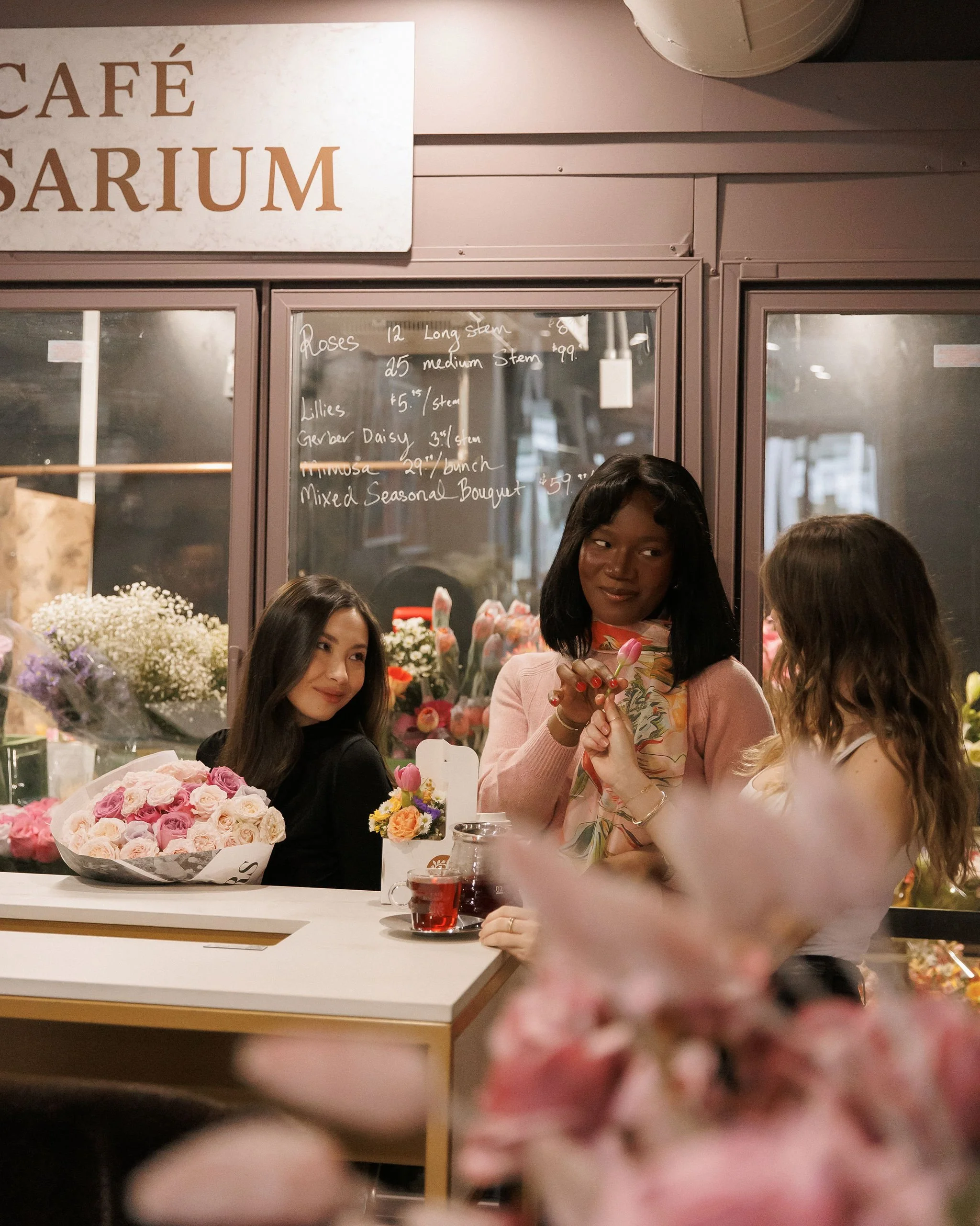 Three women in a flower shop, engaging in conversation while holding and examining flowers. One woman is sitting behind the counter with a bouquet of pink and cream roses, while the other two are standing nearby. A blackboard with handwritten plant p