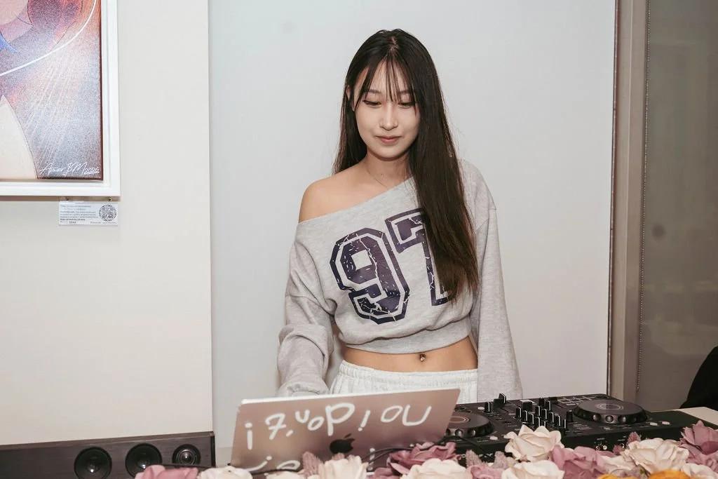 A young woman with long dark hair stands behind a DJ setup with turntables and a mixer, looking down at a sheet of paper with handwritten notes, in a room with a white wall, a framed poster, and pink flowers in the foreground.