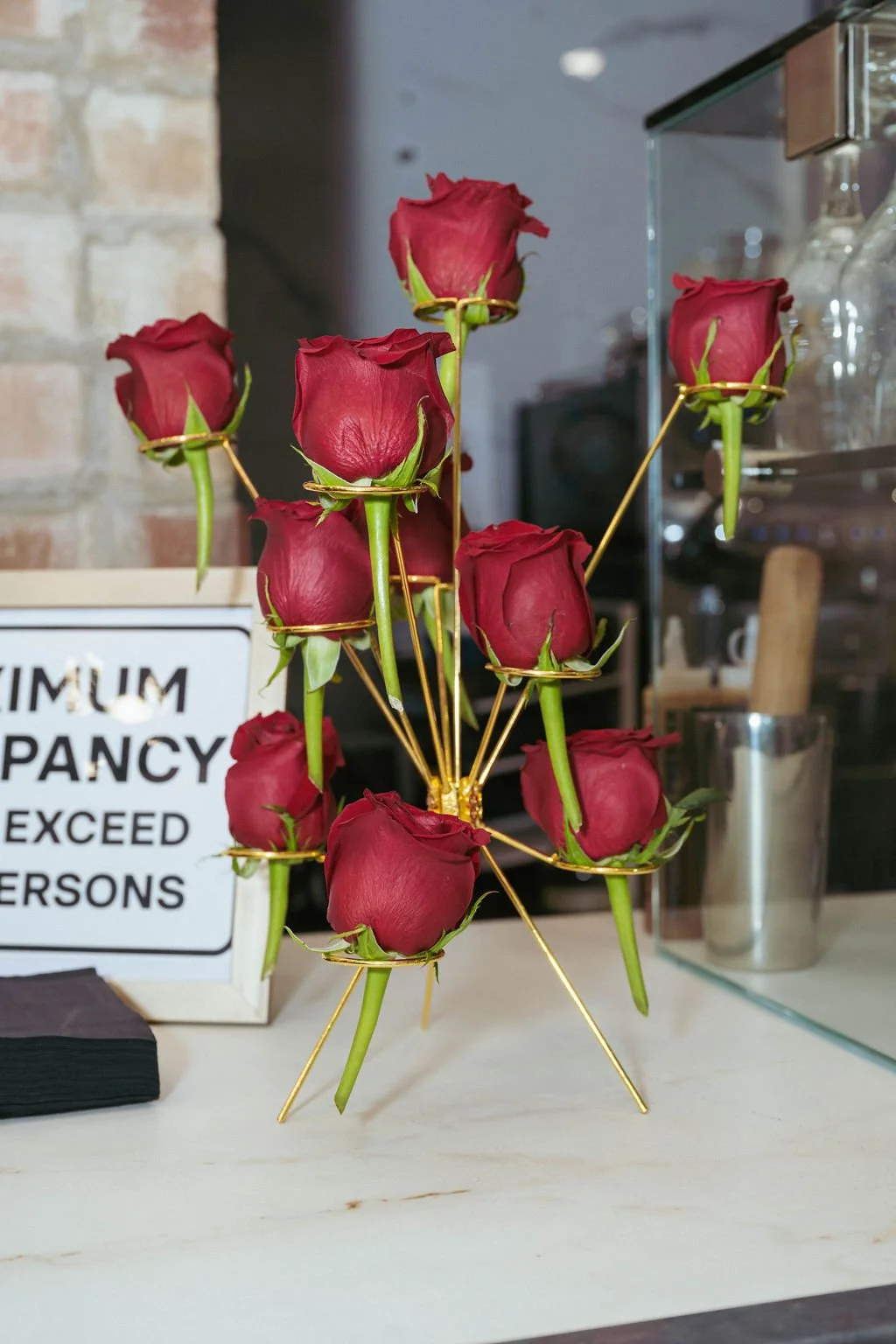 A decorative flower arrangement of red roses arranged in a gold wire stand on a white surface, with a sign and kitchen items in the background.
