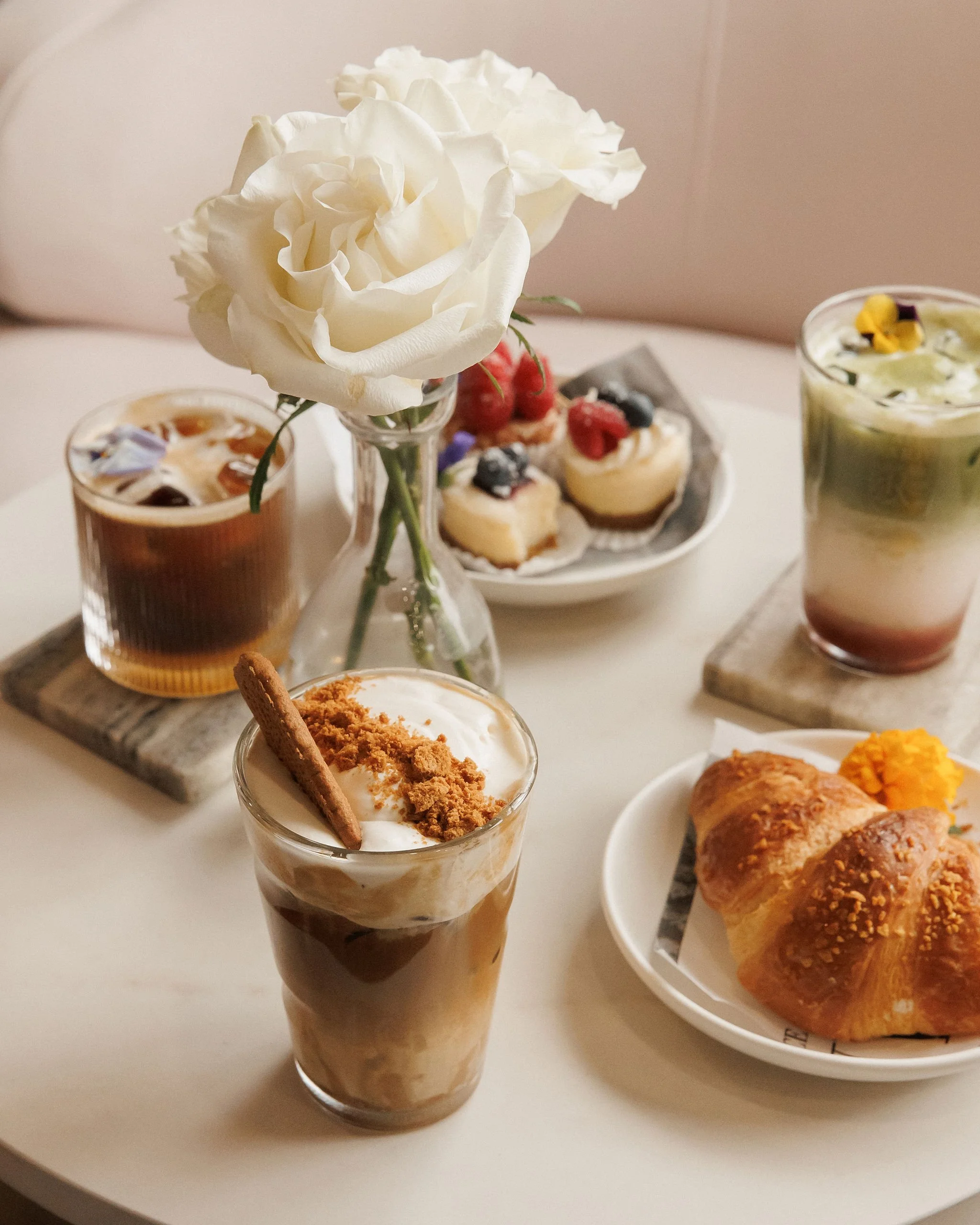A table with desserts and drinks, including a white flower in a vase, a caramel frappuccino, a berry pastry, a layered iced coffee, a croissant with a small side of orange jelly, and a plate of mini cheesecakes garnished with berries.