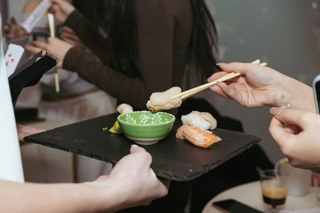 A person holding a black slate serving tray with sushi. A green bowl and sushi pieces are on the tray, and someone is using chopsticks to pick up sushi.