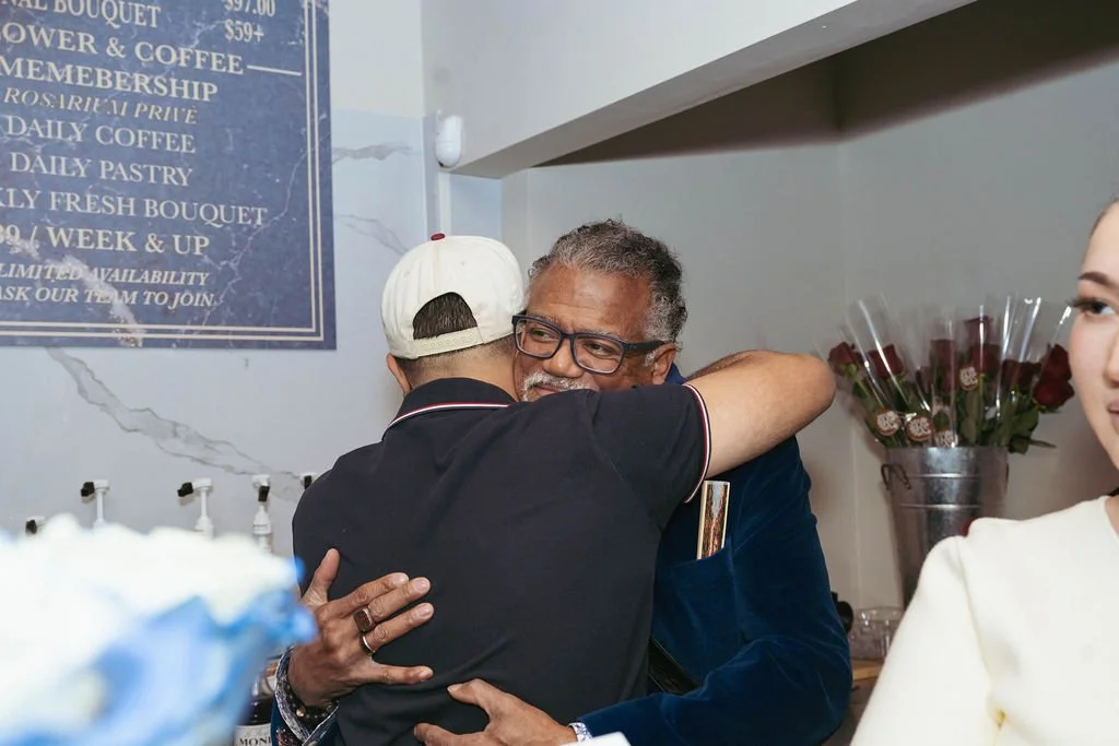 Two men hugging in a friendly embrace inside a café or restaurant, with a woman standing nearby and red roses in a vase in the background.