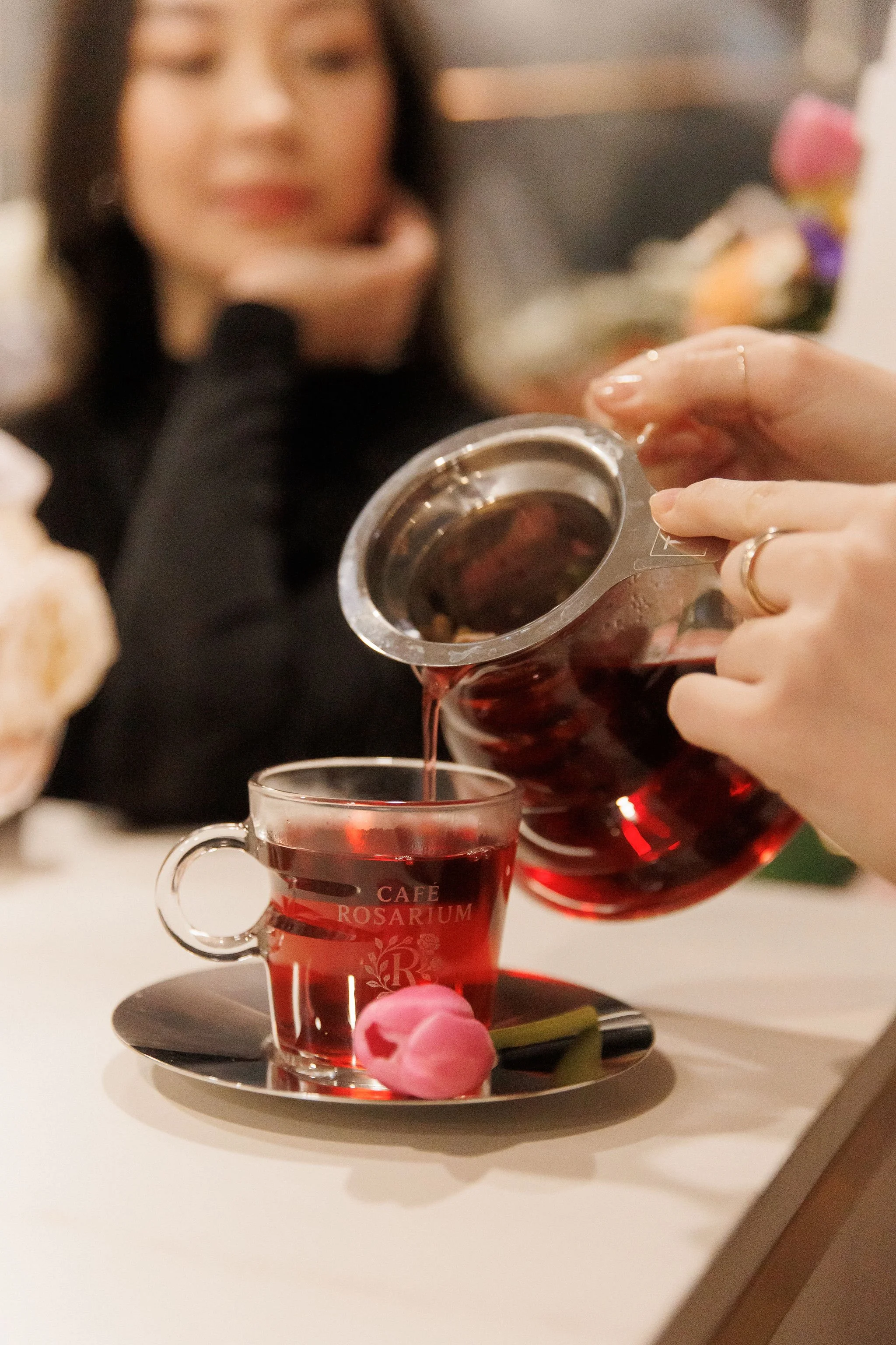 A person pouring tea from a glass teapot into a glass cup with pink flowers on a saucer, at Café Rosarium.
