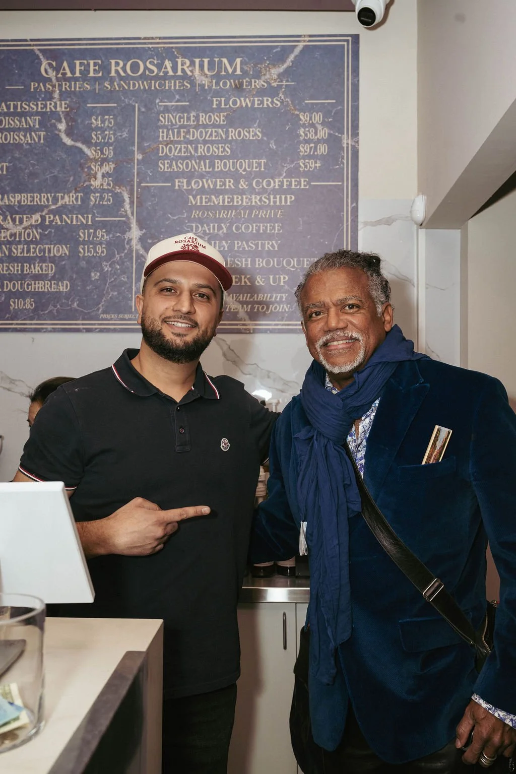 Two men smiling and posing together at Cafe Rosarium, with a menu board in the background listing pastries, flowers, and coffee.