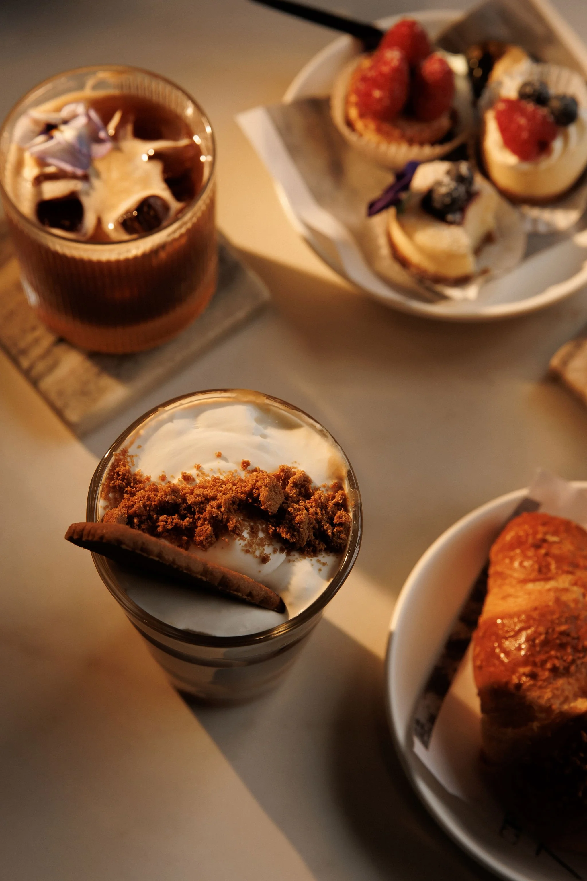 A table with a glass of vanilla milkshake topped with crushed cookies and a cookie, a glass of iced coffee with ice cubes, a plate of assorted cupcakes with berries, and a slice of layered cake.